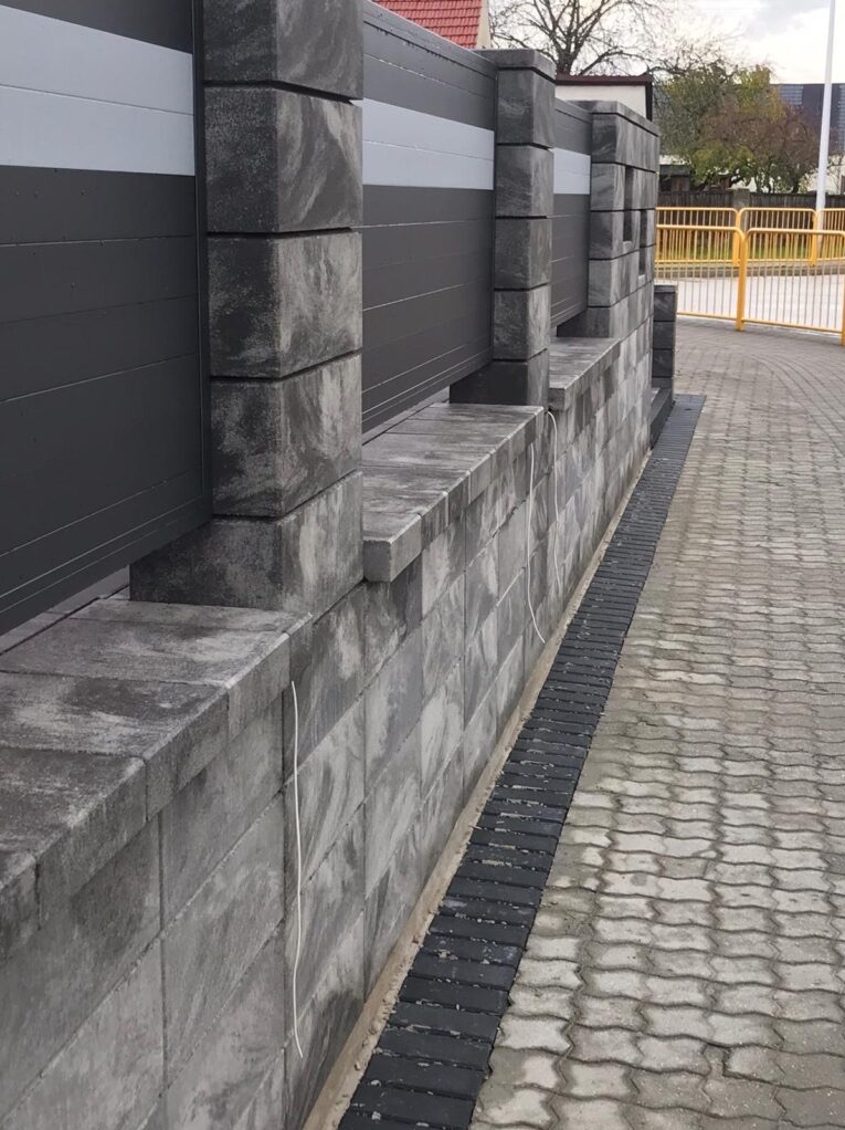 Nowoczesny mur z szarej kostki łupanej, połączony z metalowym ogrodzeniem z poziomymi panelami w odcieniach szarości i bieli, obok ułożona kostka brukowa w zygzak. Widoczne białe kable wystające ze...