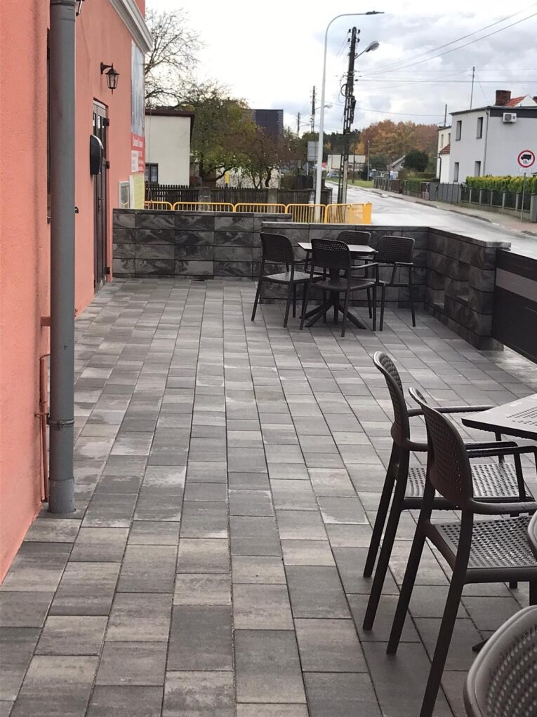 Szary chodnik z kostki brukowej przed budynkiem o łososiowej elewacji, z widocznymi stolikami i krzesłami na zewnątrz.