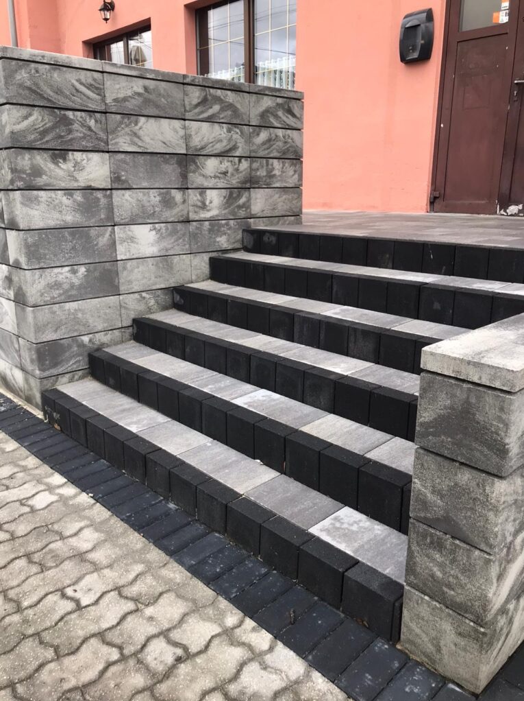 Schody wejściowe wyłożone dwukolorową kostką brukową (szary melanż i czarny) z elementami dekoracyjnymi w postaci murku z kostki o wzorze marmurkowym i obrzeżem z czarnej kostki, na tle różowej...