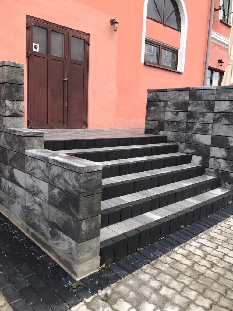 Schody wejściowe do budynku wyłożone kostką w odcieniach szarości i czerni, z widocznymi detalami faktury kostki i połączeń, na tle różowej fasady budynku z brązowymi drzwiami.