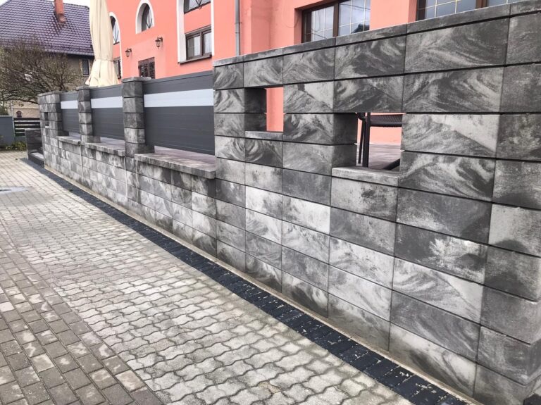 Nowoczesny mur z szarej kostki betonowej o nieregularnym wzorze, z otworami w górnej części, przylegający do chodnika z kostki brukowej i ogrodzenia z szarymi panelami.