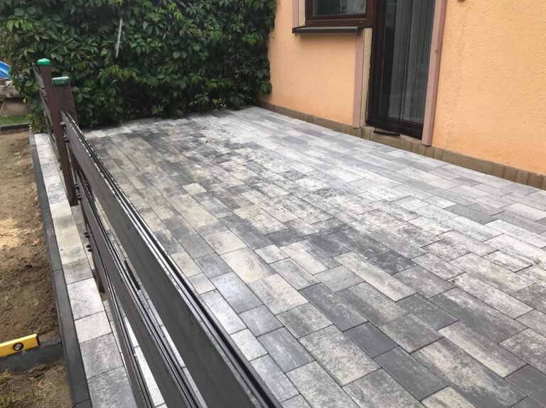 Wyłożony kostką plac z drewnianym ogrodzeniem wzdłuż budynku z oknem, widoczna poziomica na krawędzi.