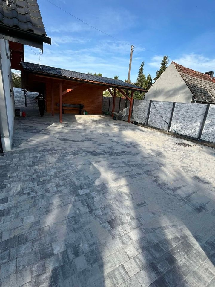 Ułożona szara kostka brukowa na posesji z drewnianą wiatą i betonowym ogrodzeniem panelowym w tle.