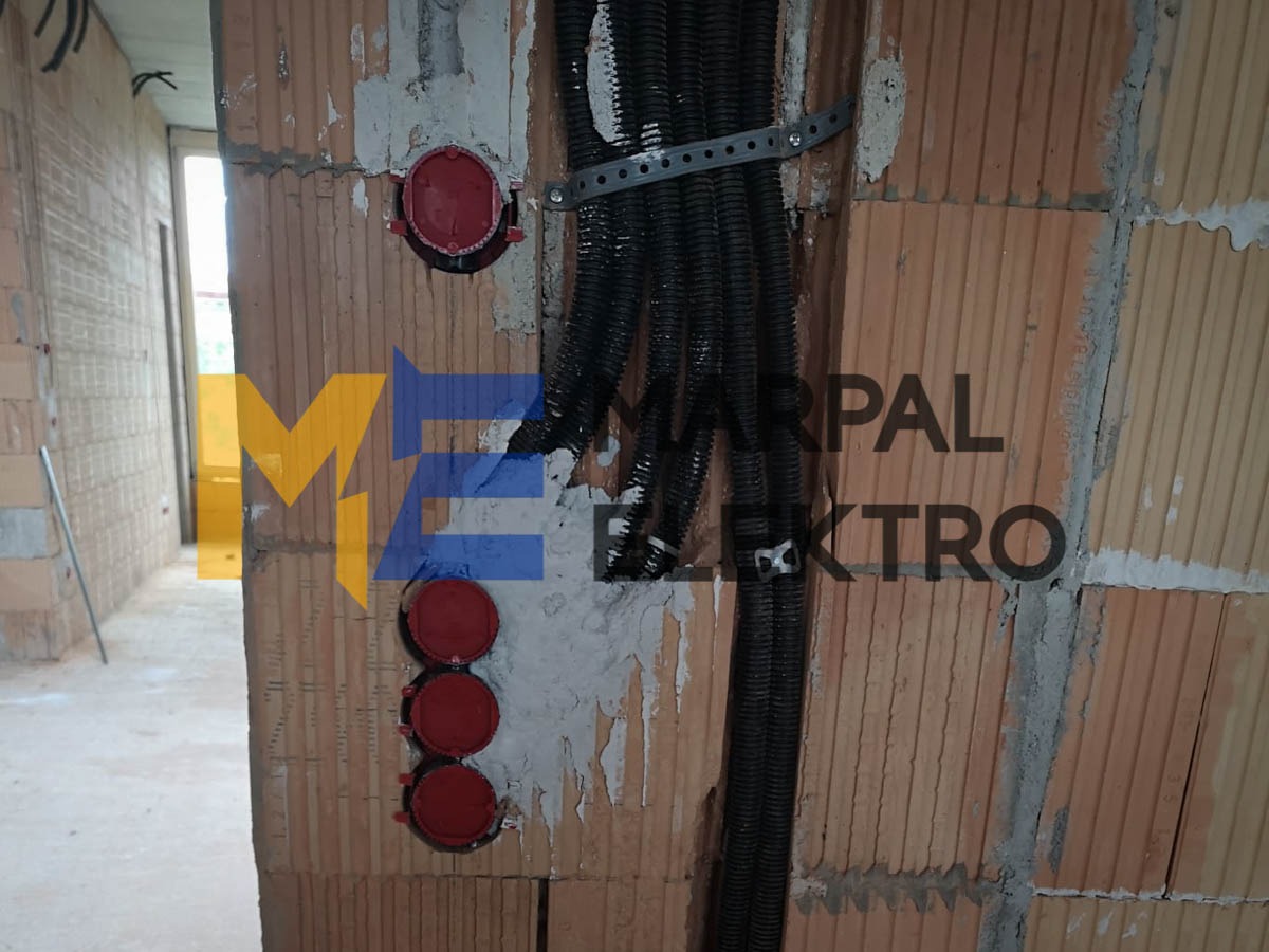 Ściana z czerwonej cegły z zamontowanymi, czarnymi rurami karbowanymi przymocowanymi metalową taśmą oraz trzema czerwonymi puszkami instalacyjnymi w pionowym rzędzie, widoczne ślady zaprawy.