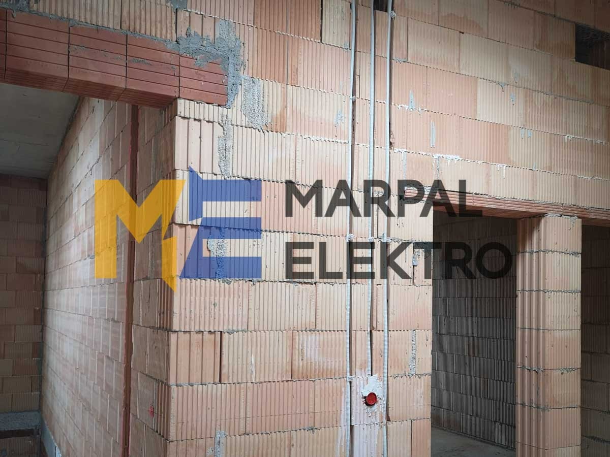Wewnętrzne ściany z pustaków ceramicznych z widocznymi przewodami elektrycznymi w peszlach, zamontowanymi natynkowo, oraz puszką instalacyjną. Na ścianie widoczne logo firmy Marpal Elektro.