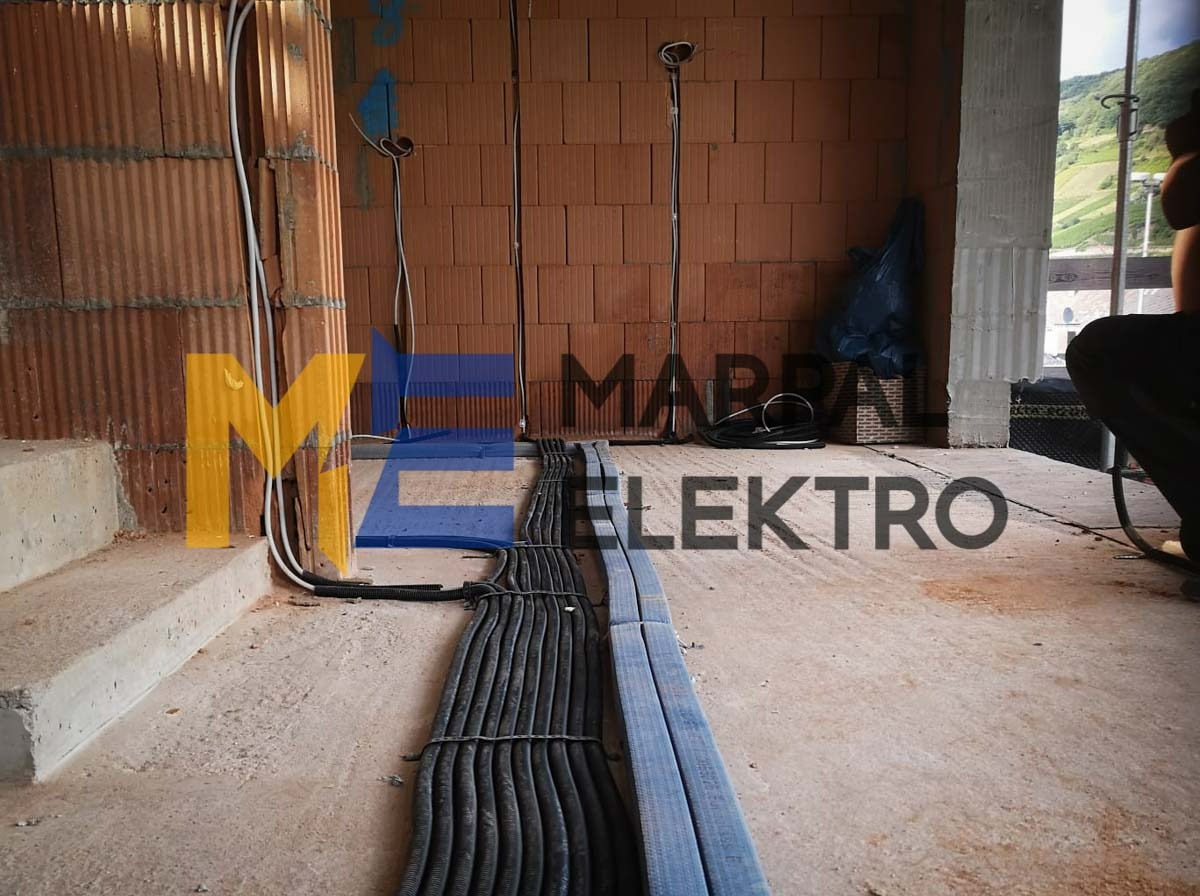 Wiązki czarnych i niebieskich kabli elektrycznych ułożone na surowej posadzce w budynku w stanie deweloperskim, widoczne przewody wychodzące ze ściany z czerwonej cegły dziurawki, częściowo...