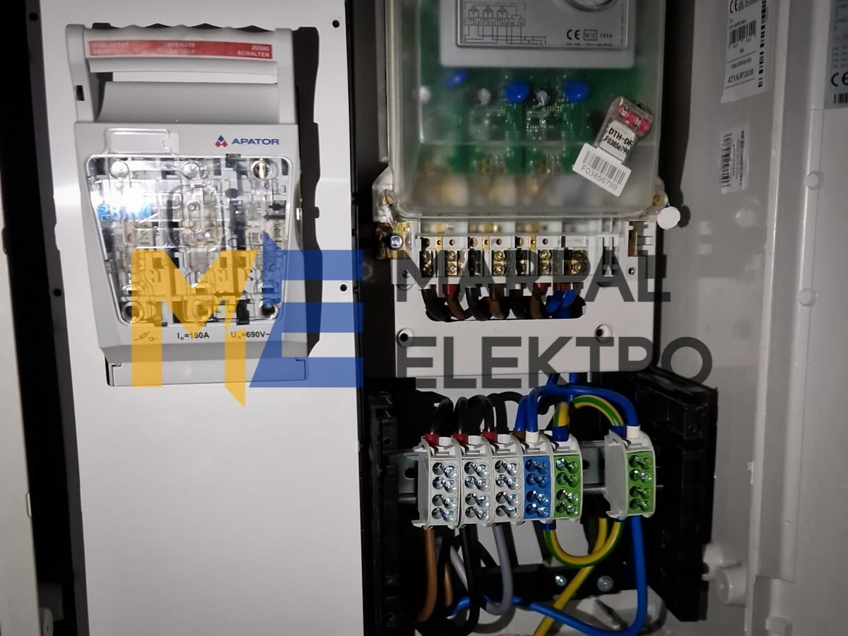 Rozdzielnica elektryczna z zainstalowanym licznikiem i zabezpieczeniami, widoczne przewody w różnych kolorach podłączone do listew zaciskowych, elementy marki Apator.
