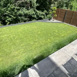 Zadbany trawnik otoczony szarą kostką brukową i dekoracyjną trawą, z drewnianym ogrodzeniem w tle, widziany z góry w słoneczny dzień.