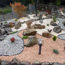 Aranżacja ogrodu z kamieniami, ozdobnymi roślinami i lampami. Widoczne różne frakcje kamieni i ścieżka. Dekoracyjny murek z kutą kratą w tle.