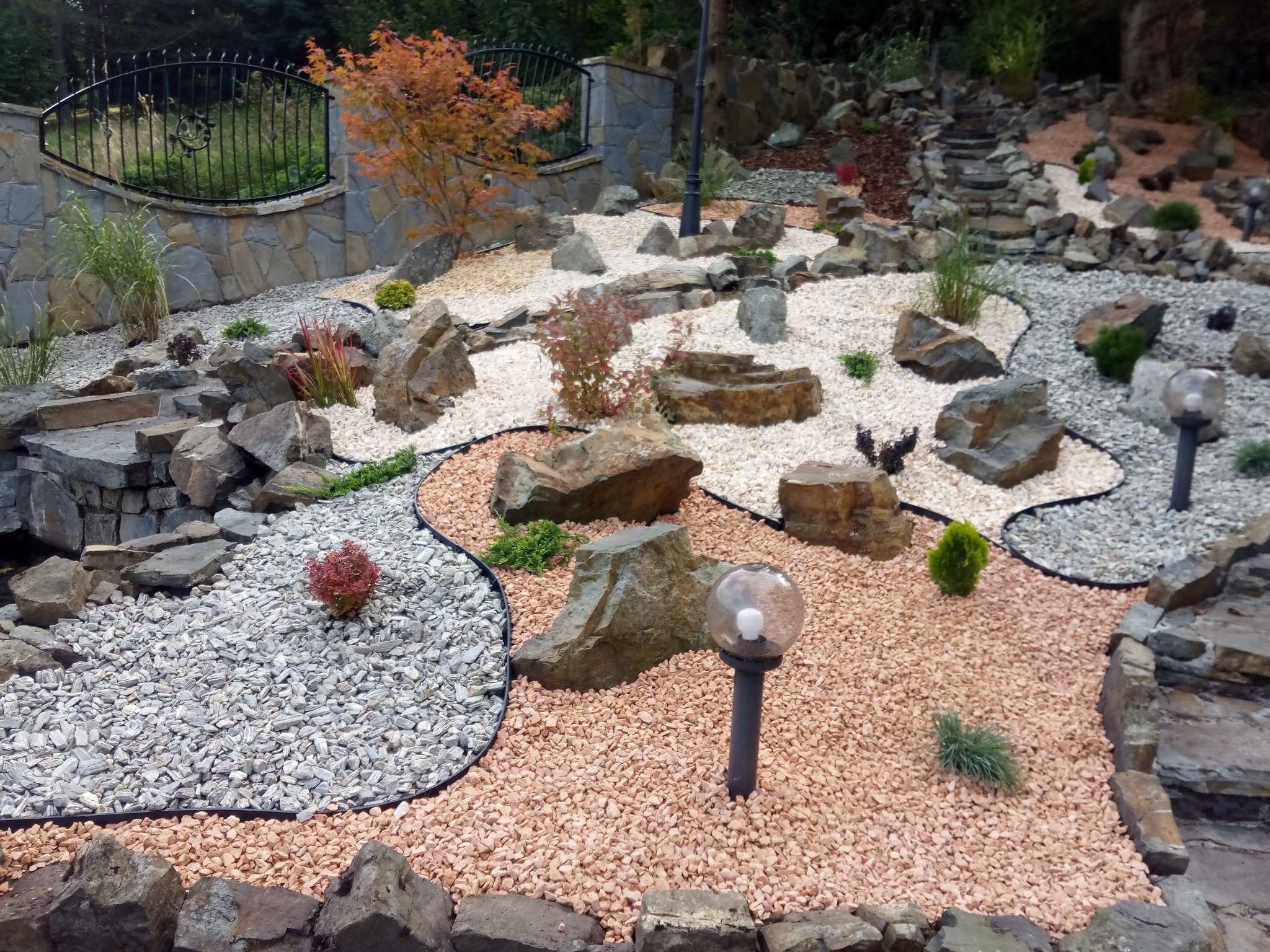 Aranżacja ogrodu z kamieniami, ozdobnymi roślinami i lampami. Widoczne różne frakcje kamieni i ścieżka. Dekoracyjny murek z kutą kratą w tle.