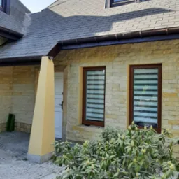 Elewacja domu z kamienia piaskowca z brązowymi oknami i szarym dachem, widoczne rolety okienne i rosnący krzew przed budynkiem.