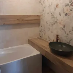 Nowoczesna łazienka z wanną, umywalką w kolorze oliwkowym na drewnianym blacie, baterią w kolorze miedzi i płytkami w kwiatowe wzory na ścianie.