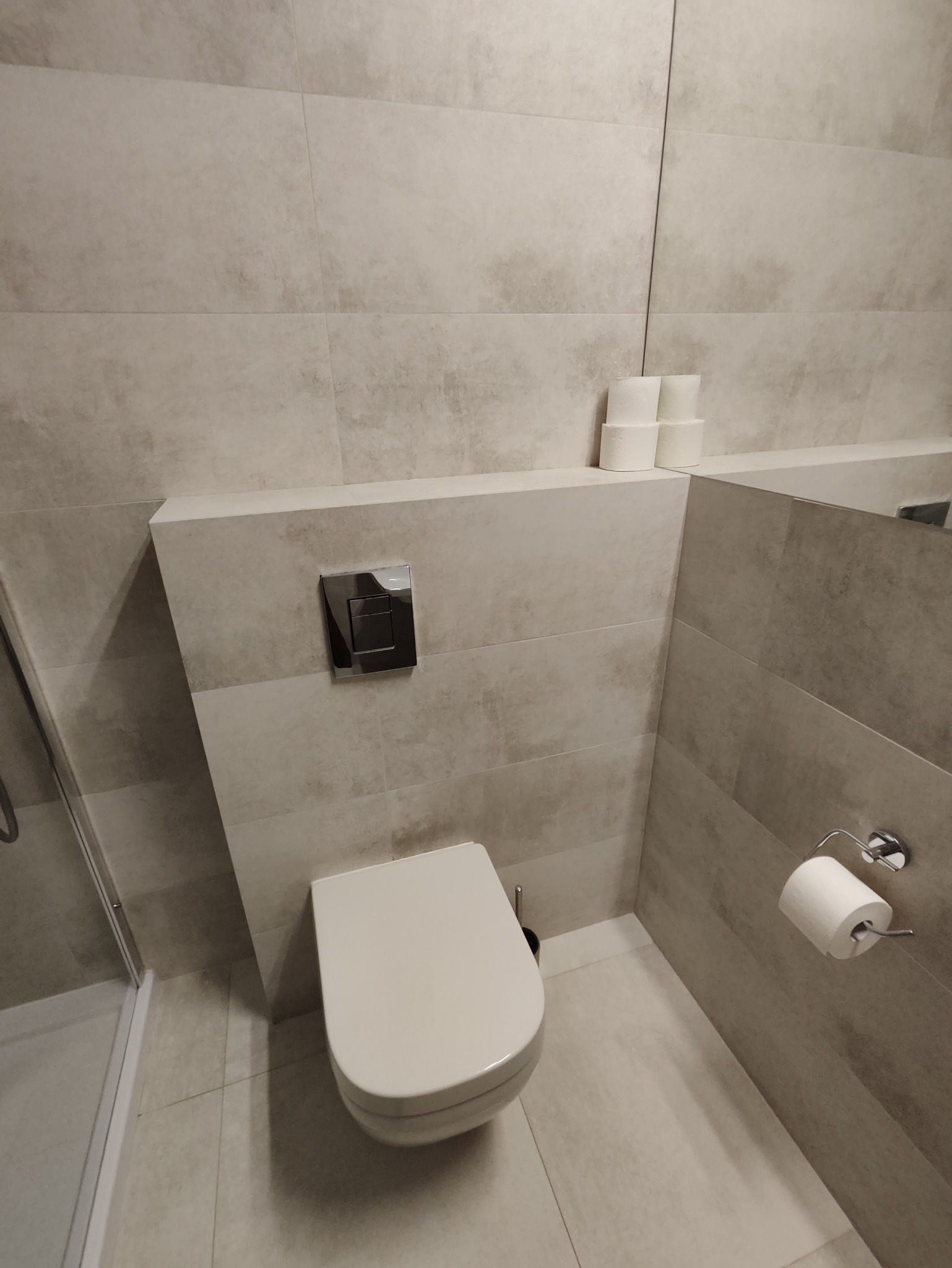 Nowoczesna toaleta podwieszana z chromowanym przyciskiem spłukującym, płytki imitujące beton, rolka papieru toaletowego na chromowanym uchwycie i zapas papieru na półce w tle.