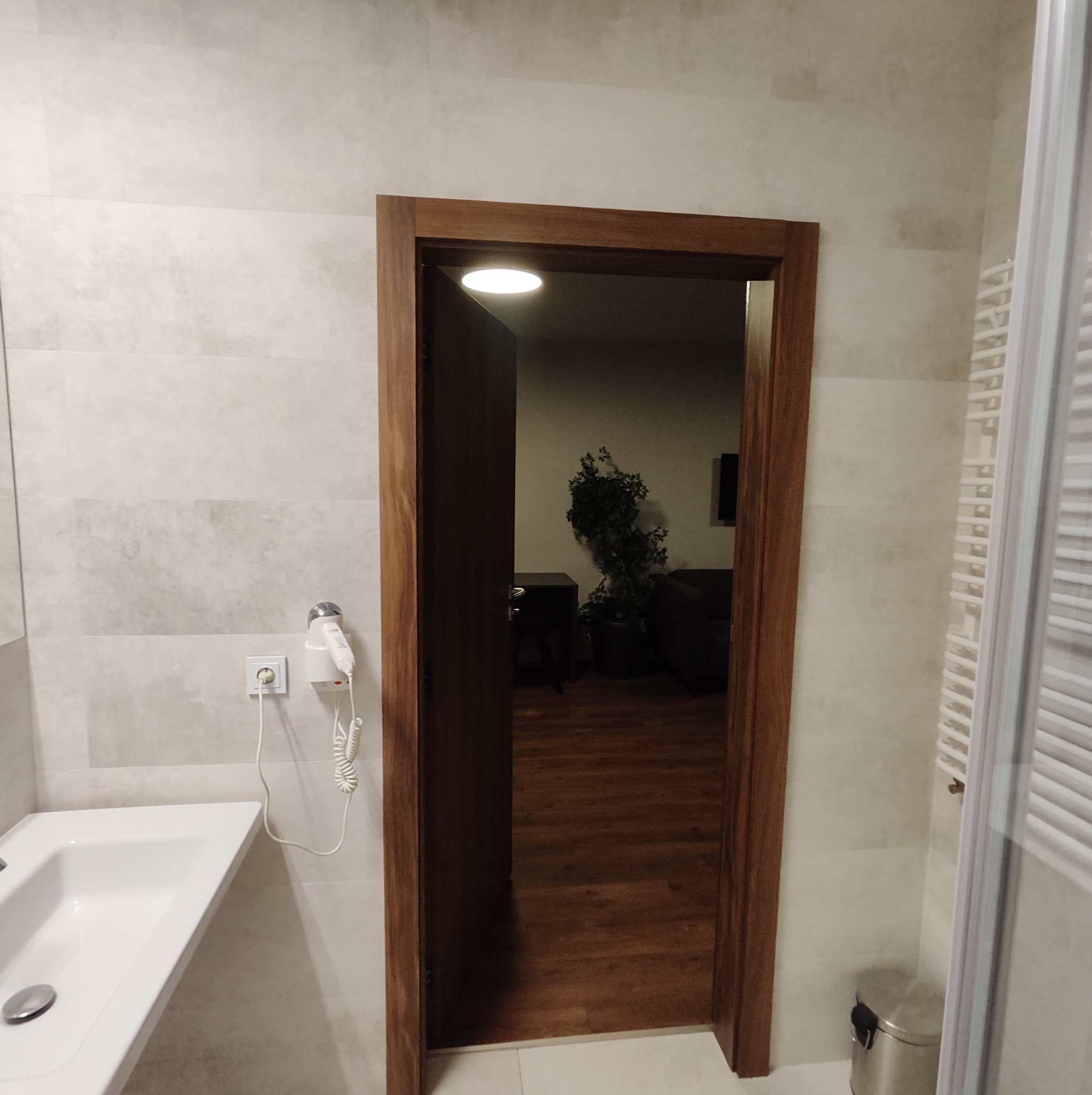 Nowoczesna łazienka z umywalką, grzejnikiem drabinkowym i otwartymi drewnianymi drzwiami do pokoju z rośliną doniczkową i ciemnymi meblami.