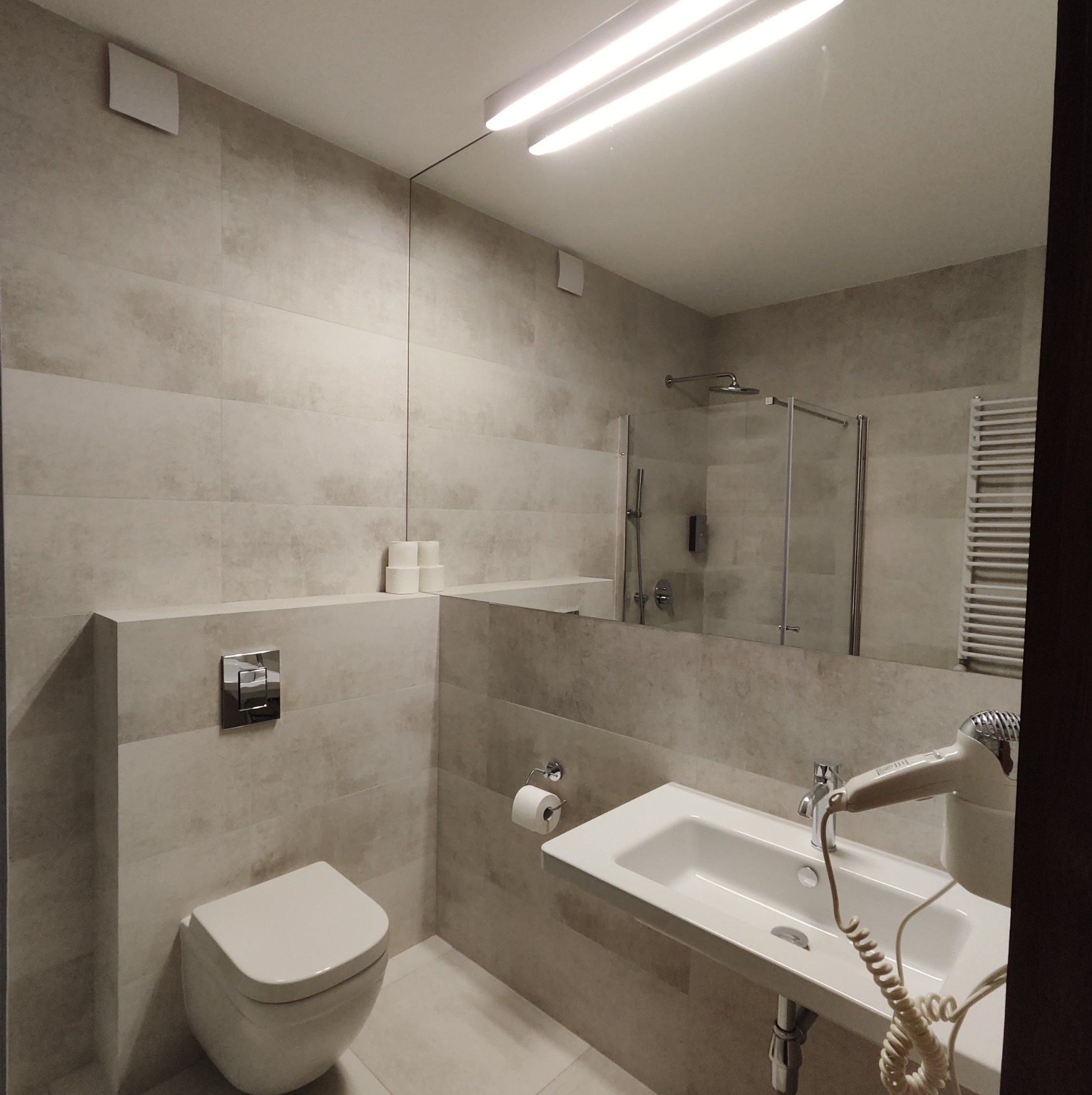 Nowoczesna łazienka z beżowymi płytkami, podwieszaną toaletą z chromowanym przyciskiem spłukiwania, prostokątną umywalką na stelażu i dużym lustrem, w którym odbija się kabina prysznicowa...