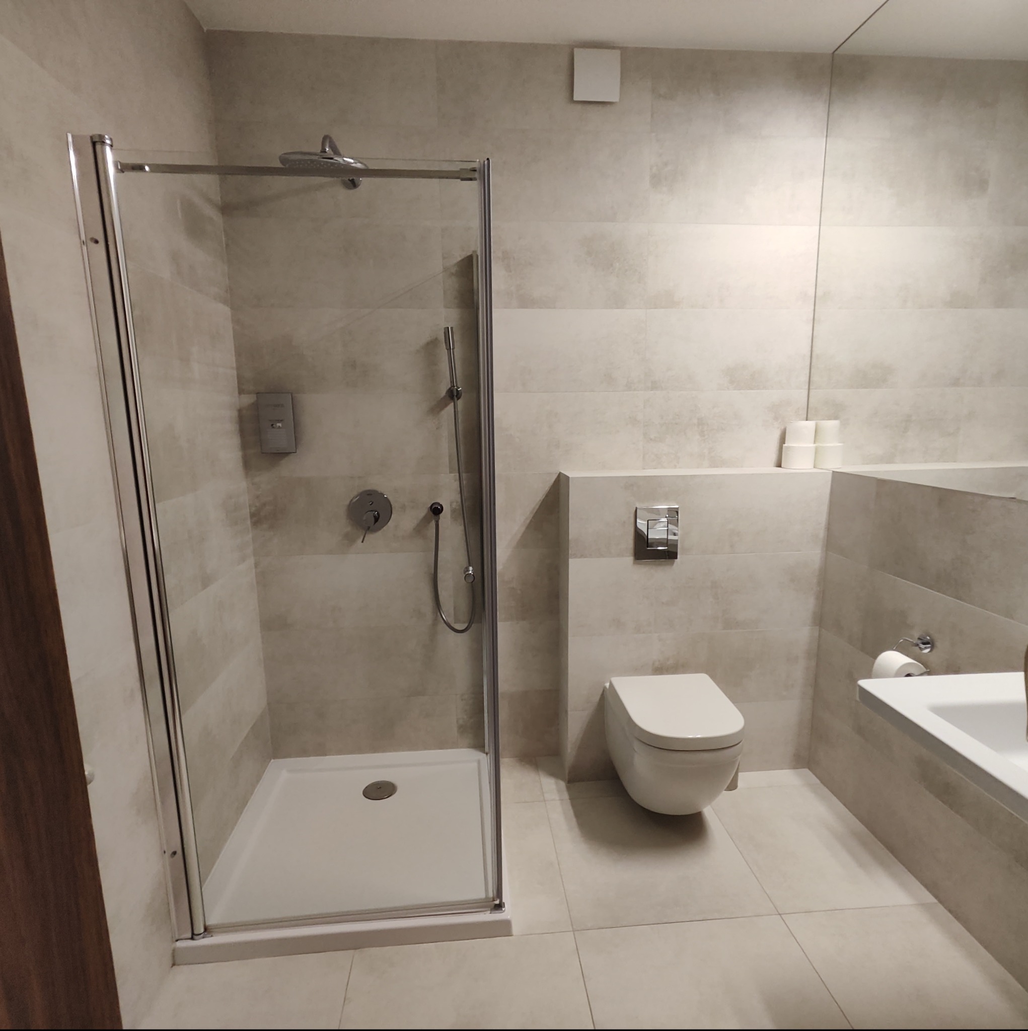 Nowoczesna łazienka w odcieniach beżu z kabiną prysznicową ze srebrnymi ramami, podwieszaną toaletą i lustrem odbijającym rolki papieru toaletowego.