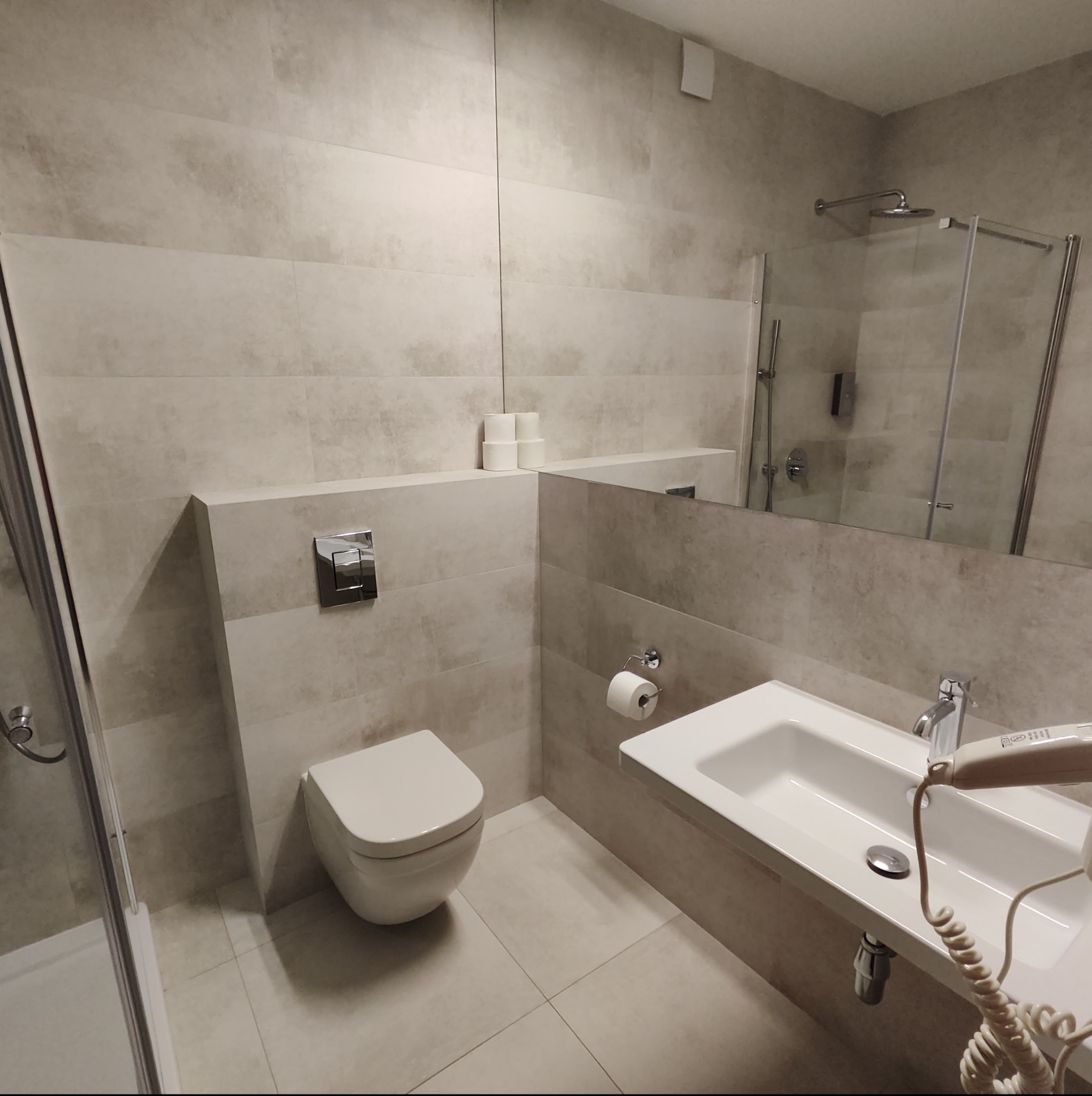 Wykończona łazienka w odcieniach beżu w Łodzi, z wiszącą toaletą, umywalką z baterią i kabiną prysznicową z chromowanymi detalami. Na ścianach widoczne płytki imitujące kamień.