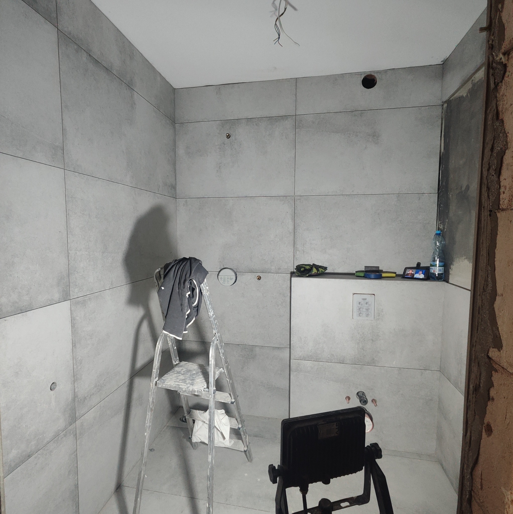 Szara łazienka w trakcie remontu: ułożone płytki, drabina z ubraniem, lampa budowlana, niedokończone ściany.