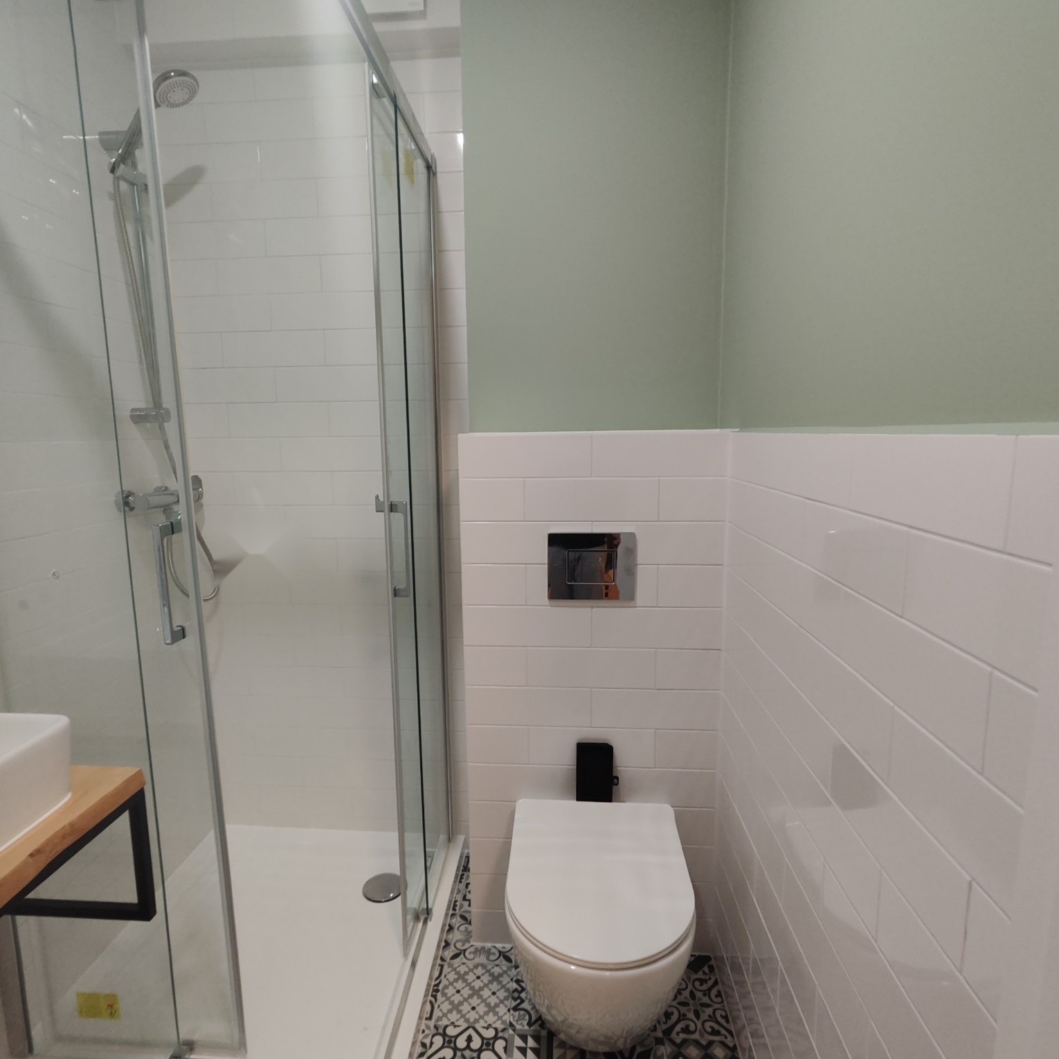 Nowoczesna łazienka z kabiną prysznicową, wiszącą toaletą i białymi płytkami ułożonymi w cegiełkę, akcentem jest podłoga w czarno-białe wzory geometryczne oraz ściana pomalowana na pistacjowy kolor.