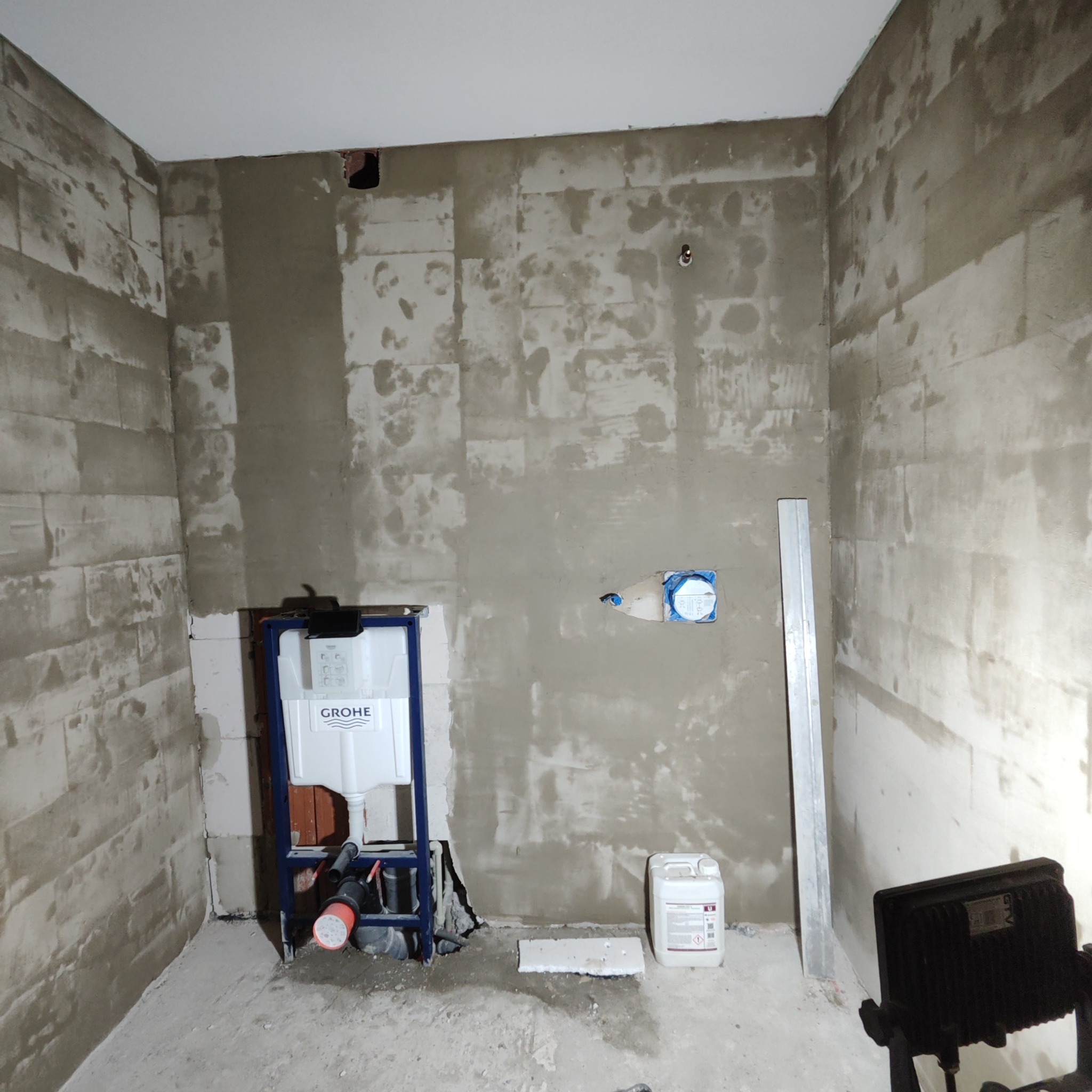 Wnętrze łazienki w trakcie prac wykończeniowych, widoczne ściany z częściowo nałożoną zaprawą, stelaż podtynkowy WC marki Grohe, pojemnik z płynem gruntującym oraz lampa budowlana.