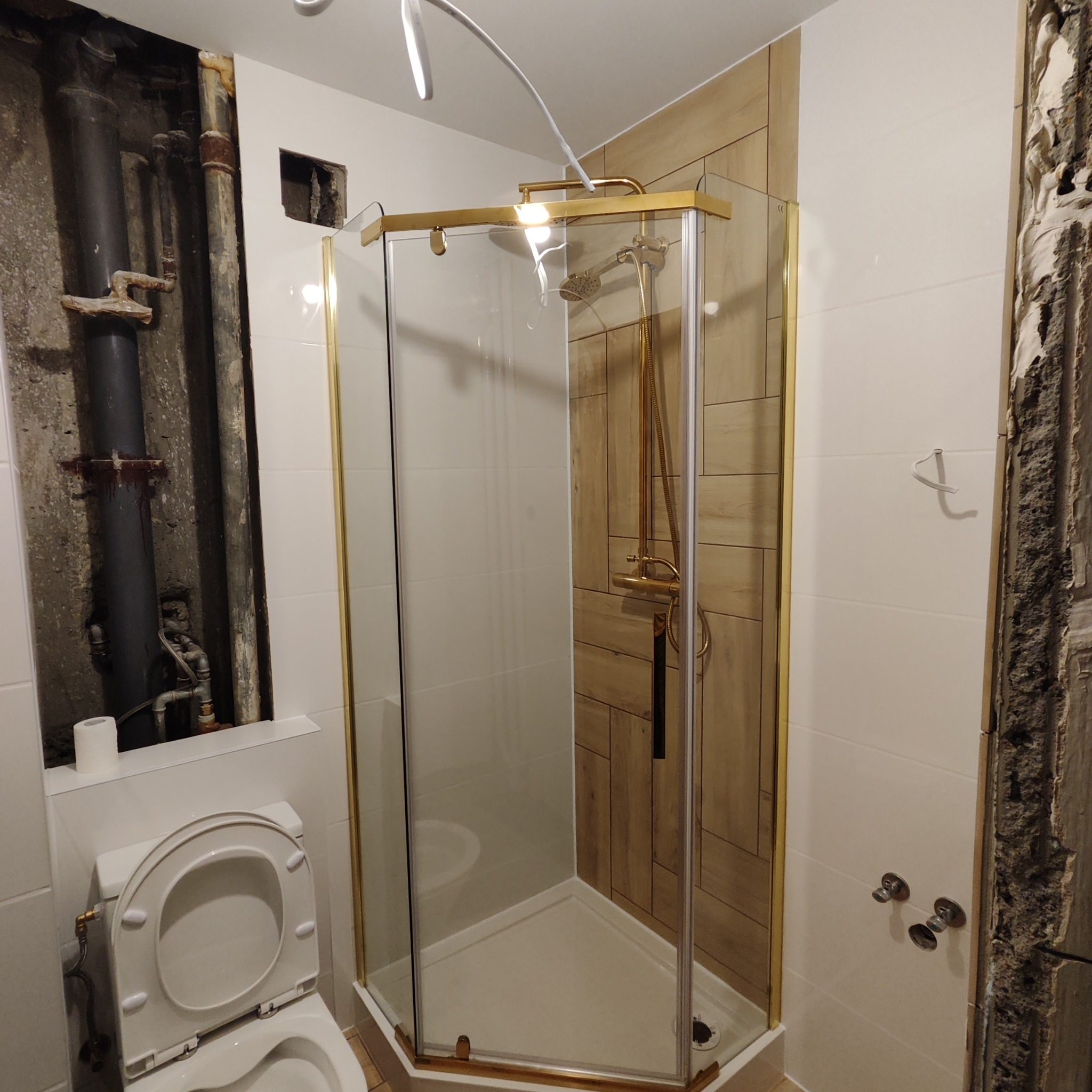 Narożna kabina prysznicowa ze złotymi profilami w trakcie montażu w łazience, widoczne niedokończone prace wykończeniowe, odsłonięte rury i instalacje.
