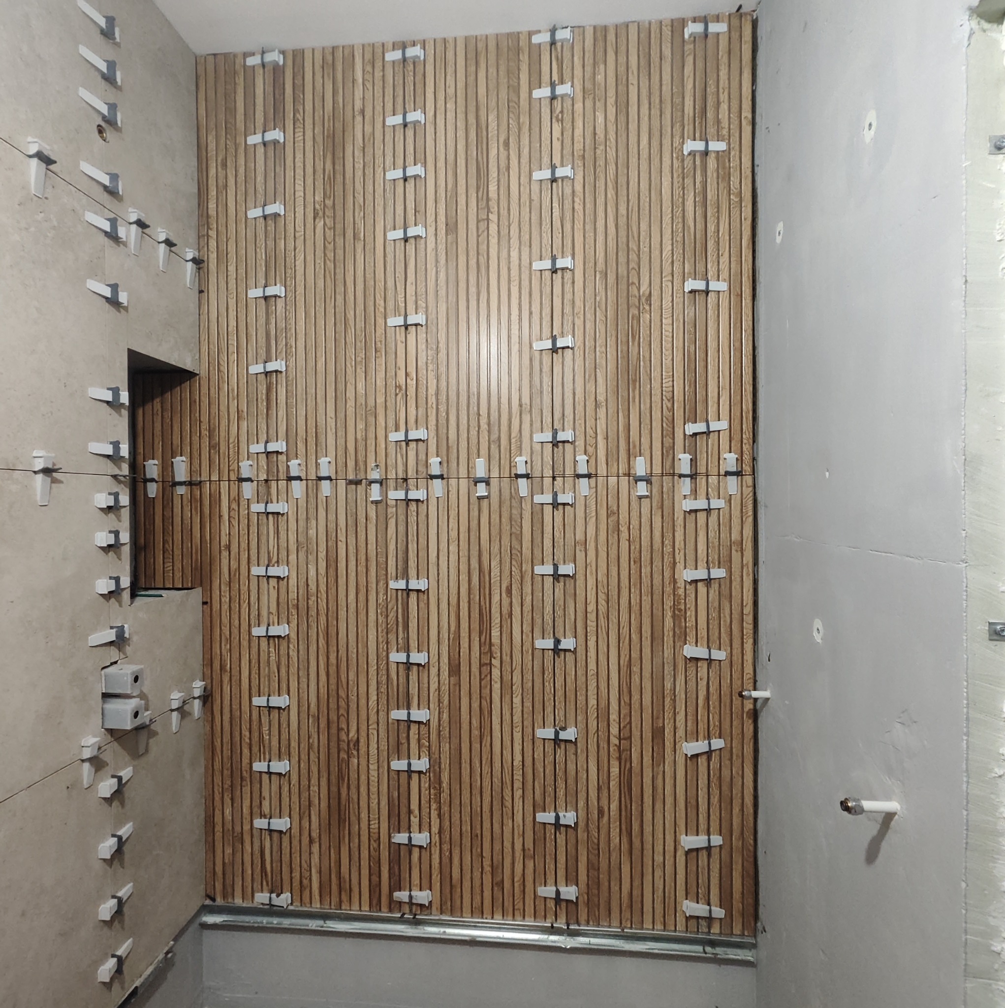 Montaż drewnopodobnych paneli ściennych w łazience, widoczne klipsy poziomujące i pionujące, surowa ściana obok.