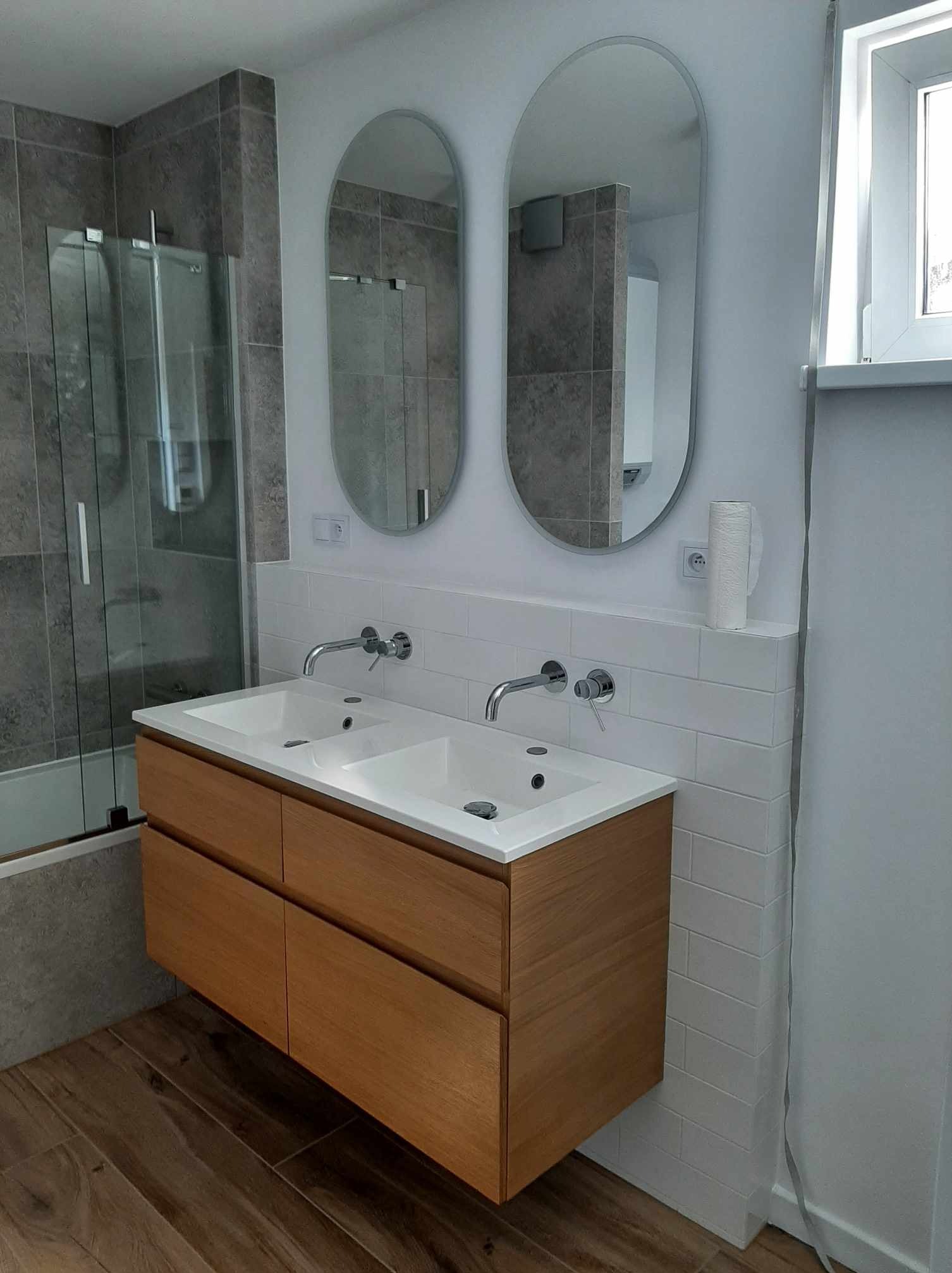 Nowoczesna łazienka z drewnianą szafką pod umywalką z dwiema owalnymi umywalkami, dwoma owalnymi lustrami bez ram, kabiną prysznicową ze szklanymi drzwiami i drewnopodobną podłogą z płytek.