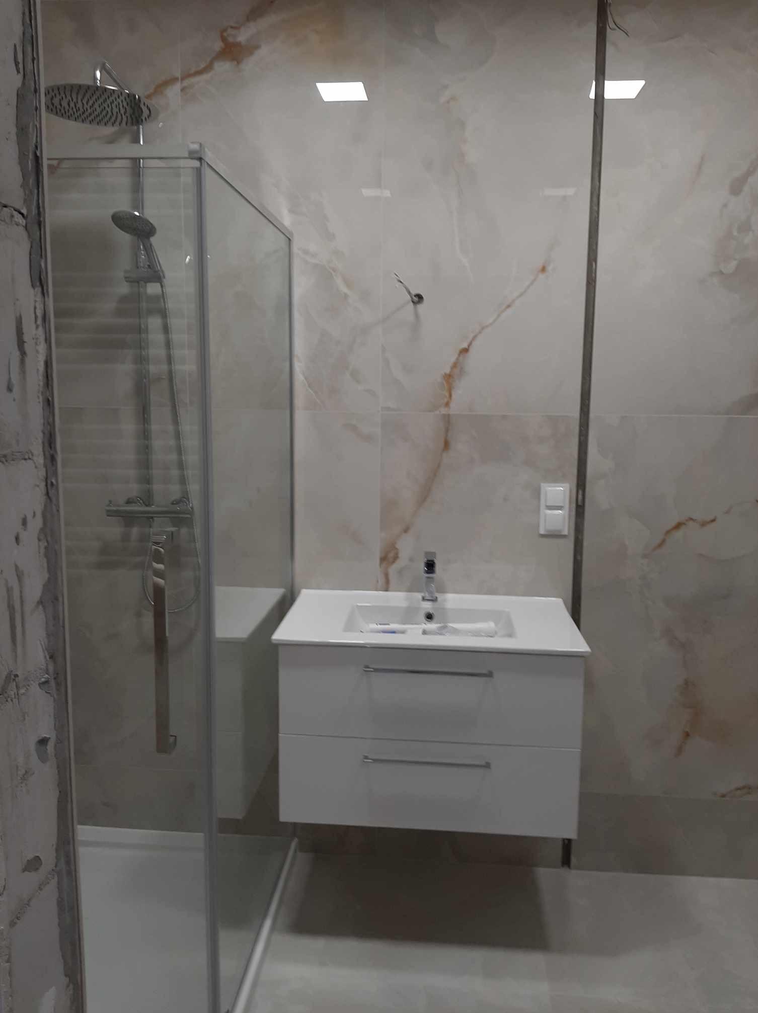 Nowoczesna łazienka w trakcie remontu: biała szafka pod umywalkę z dwiema szufladami, kabina prysznicowa ze szklanymi drzwiami i deszczownicą, płytki imitujące marmur na ścianach, widoczne ślady...