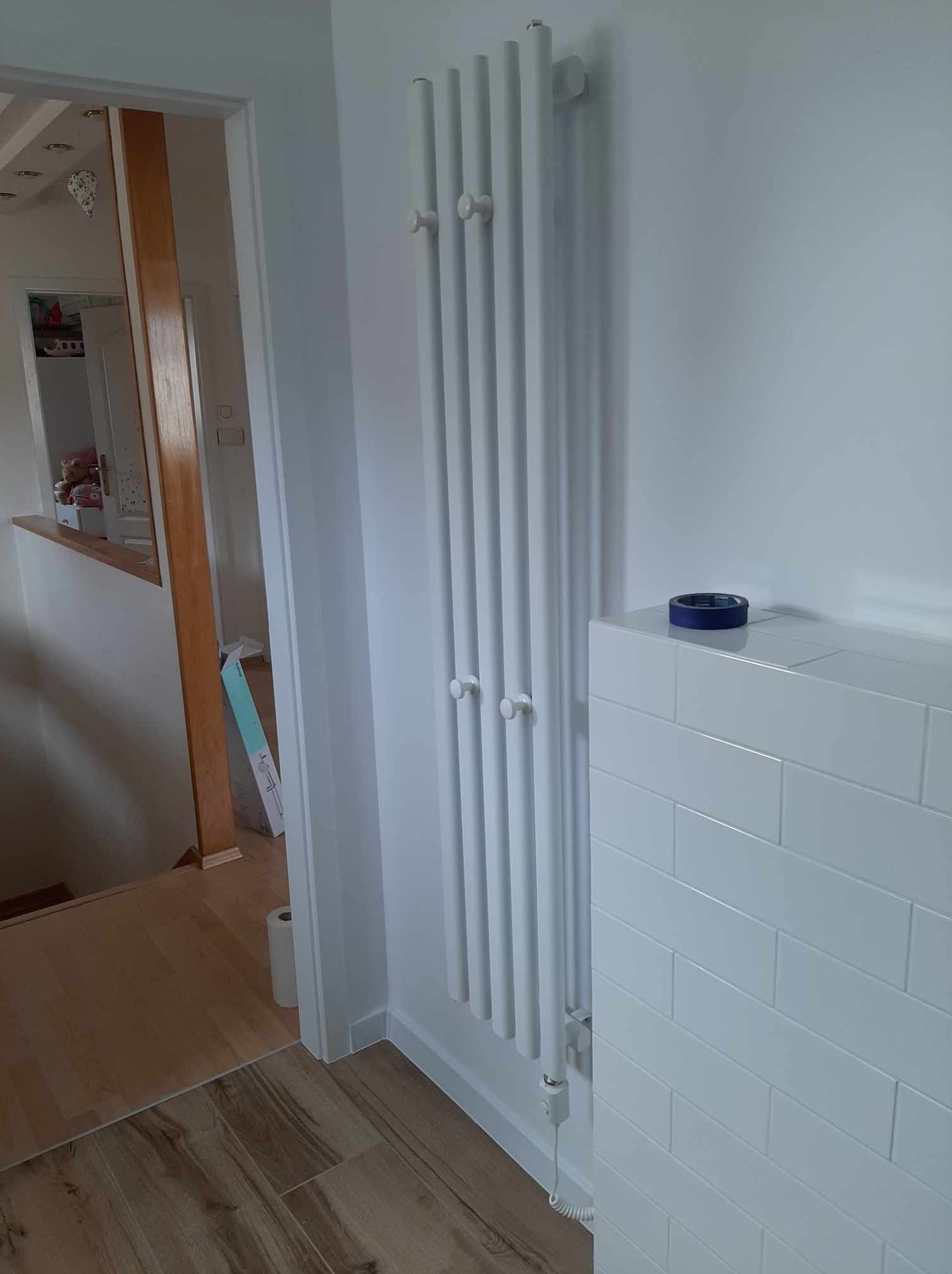 Biały, pionowy grzejnik żeberkowy z haczykami, zamontowany na ścianie wyłożonej białymi, prostokątnymi płytkami, z niebieską taśmą malarską na górze; w tle widoczne drewniane elementy wykończeniowe...