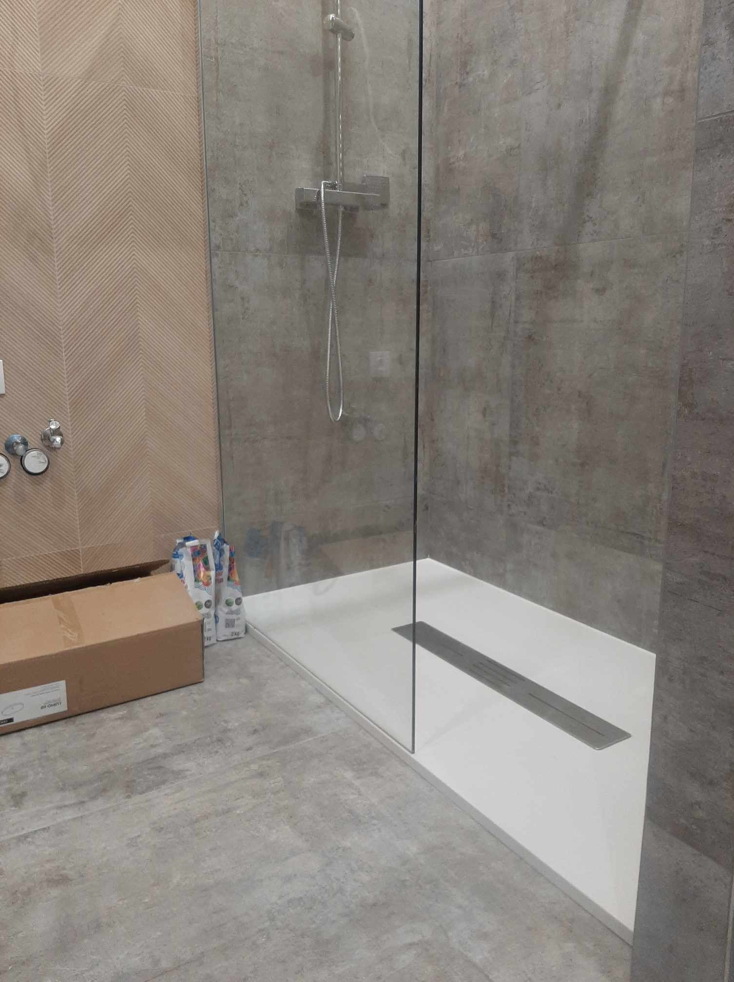Nowoczesna kabina prysznicowa z odpływem liniowym, szklana ściana, płytki imitujące kamień, pudełko kartonowe i worki z zaprawą obok.