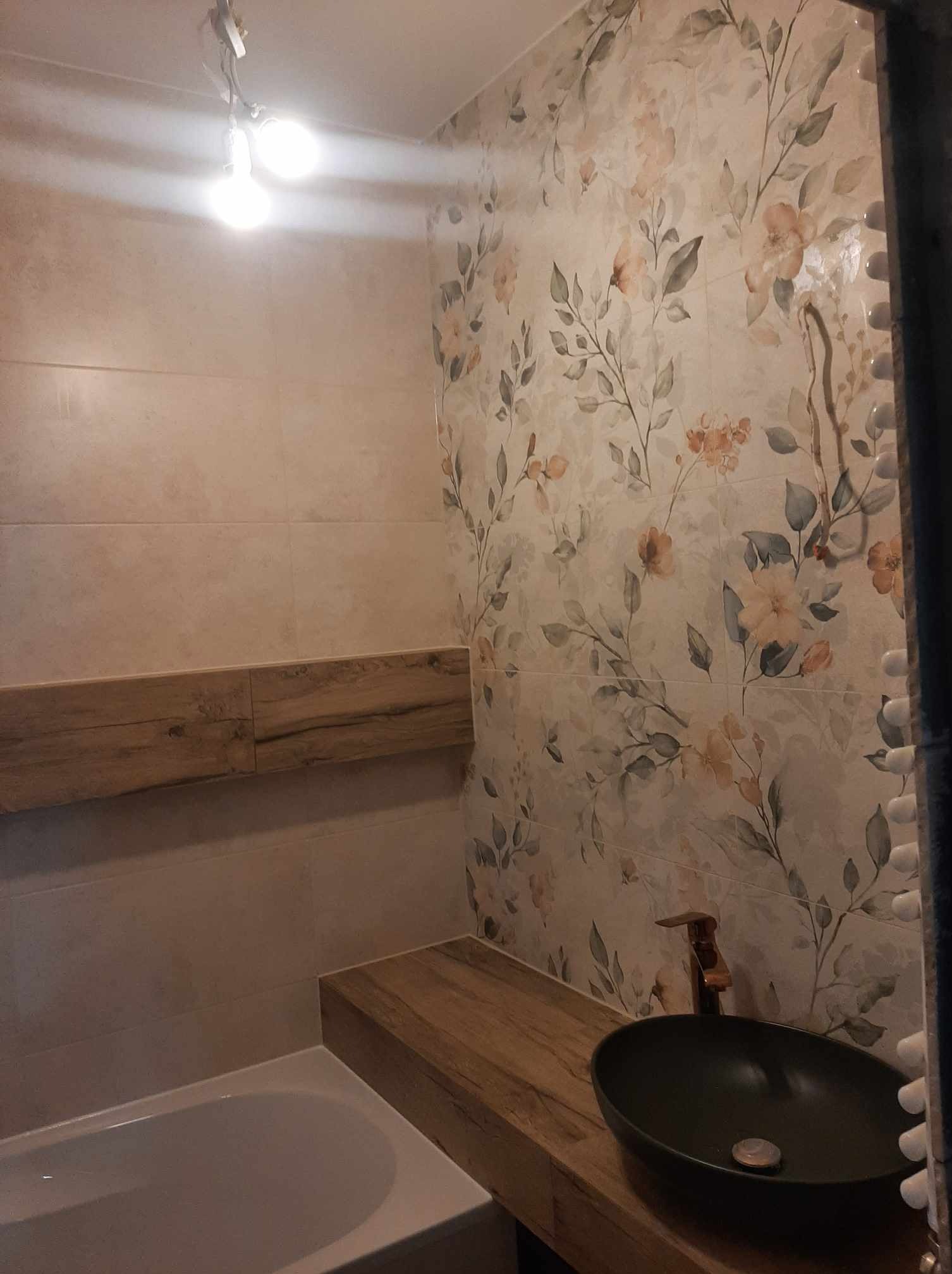 Umywalka nablatowa w kolorze ciemnej zieleni z miedzianą baterią na drewnianym blacie w łazience z płytkami w kwiatowy wzór, widoczna wanna i drewniana półka.