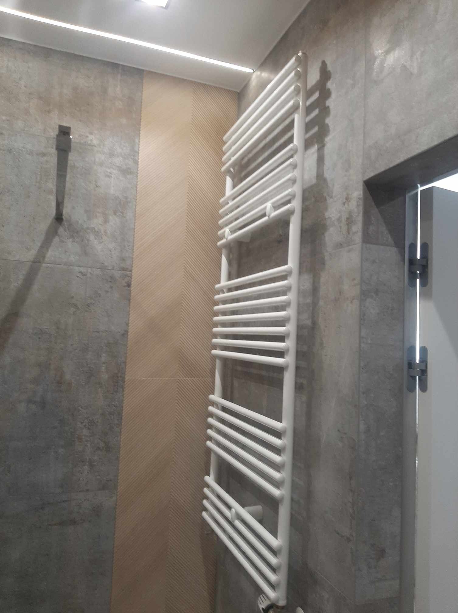Biały, drabinkowy grzejnik łazienkowy na tle szarej, betonowej ściany i beżowej ściany z delikatnym wzorem, widoczne zawiasy białych drzwi.