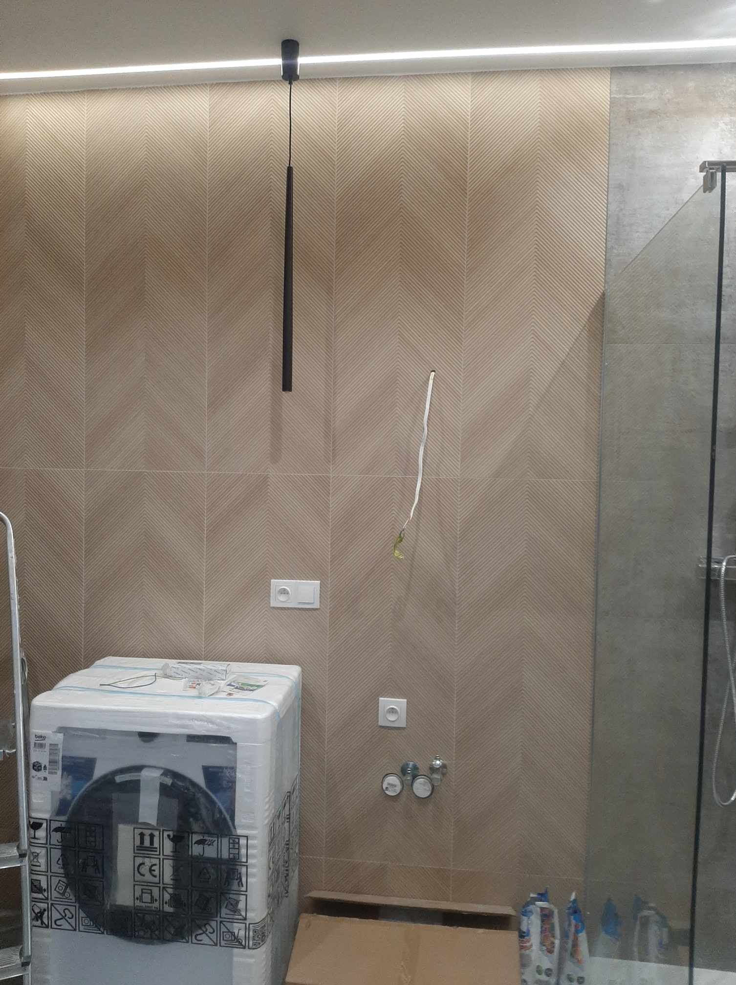 Beżowe płytki chevron na ścianie łazienki, nowa pralka w folii ochronnej, wisząca lampa i szklana kabina prysznicowa w trakcie montażu.