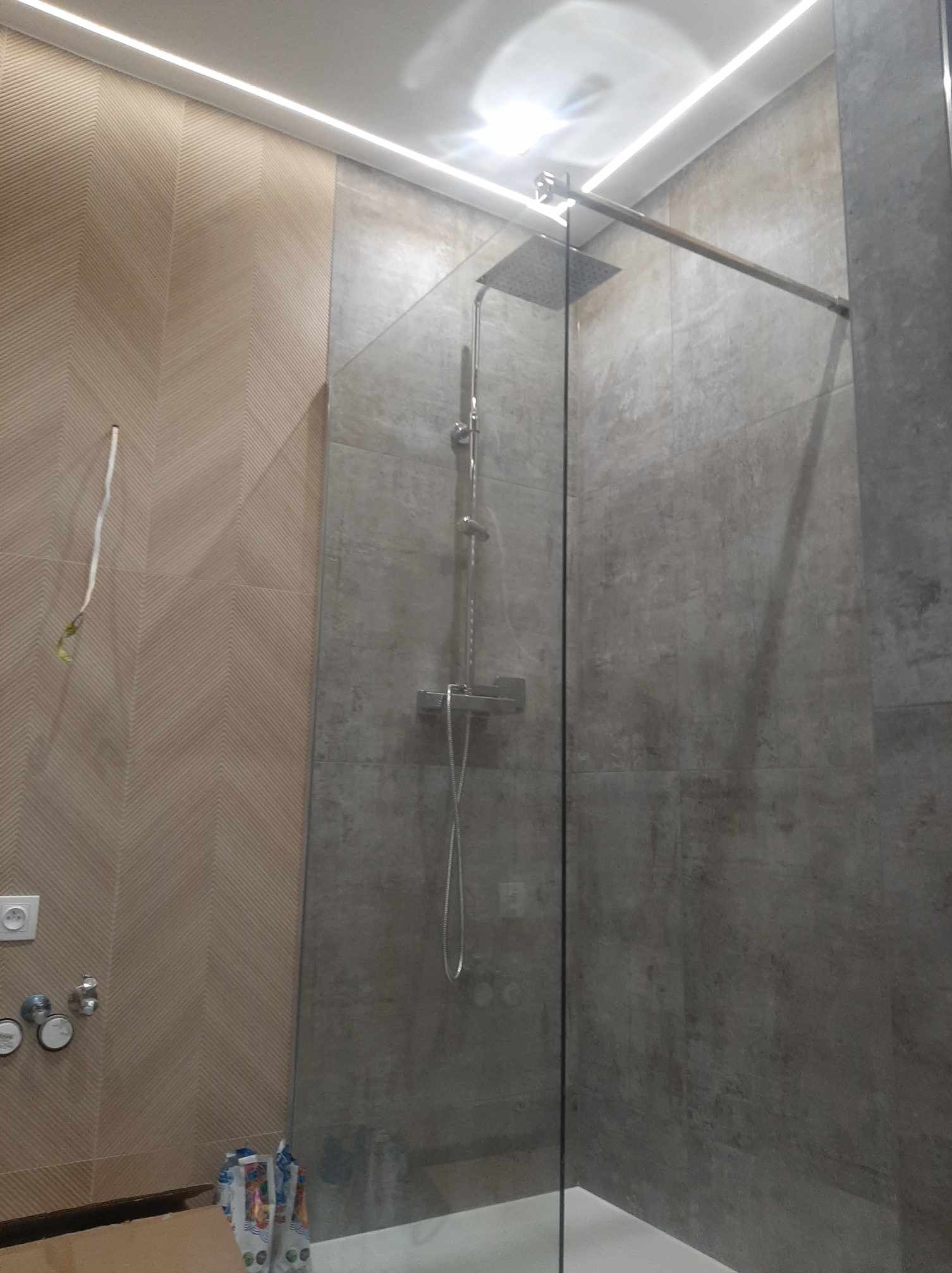 Wykończona kabina prysznicowa typu walk-in z deszczownicą, szklaną ścianką i szarą, teksturowaną okładziną ścienną, uzupełniona beżową ścianą z wzorem jodełki i listwą LED podsufitową.