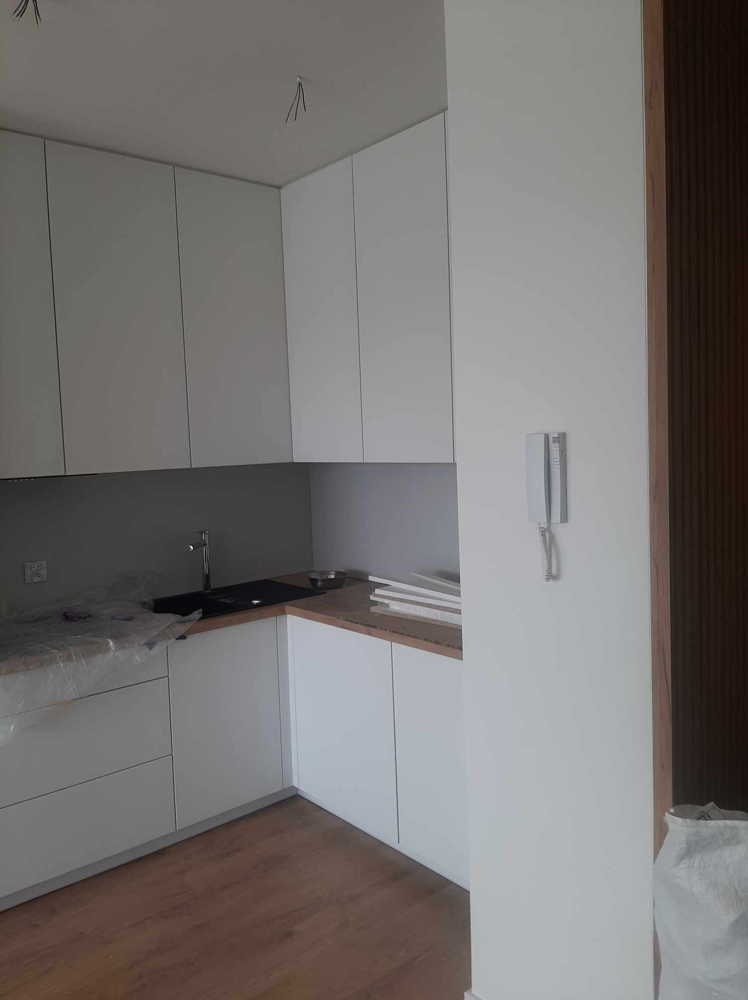 Wnętrze kuchni w trakcie wykańczania: białe szafki kuchenne, drewniany blat częściowo zakryty folią ochronną, widoczne przewody elektryczne wystające z sufitu, zamontowany domofon na ścianie.