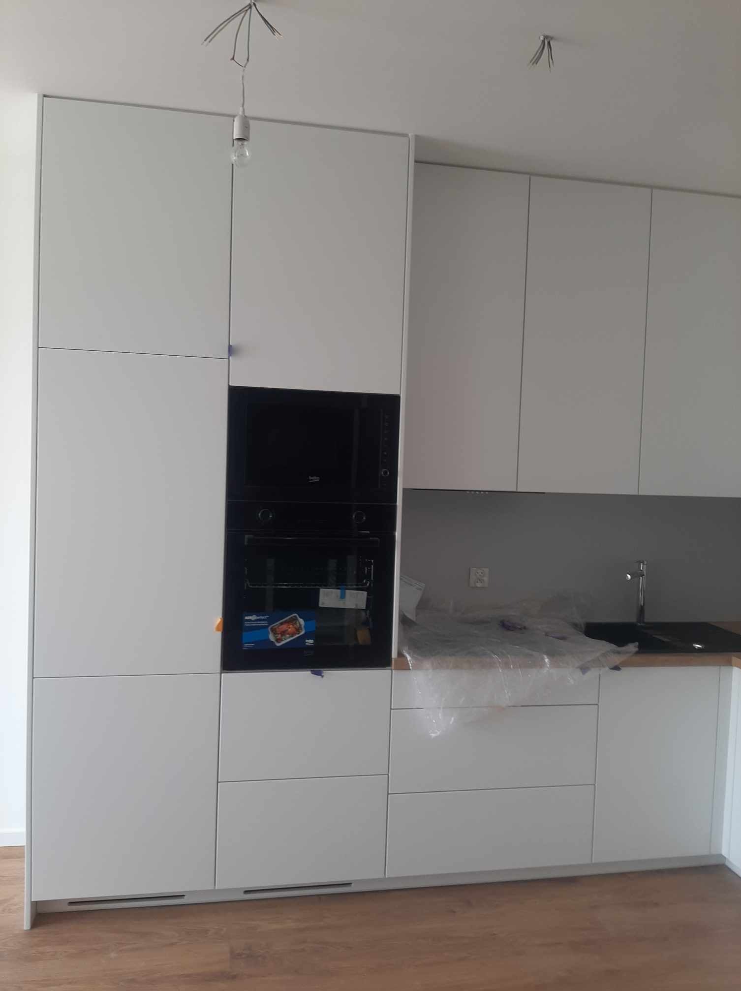 Nowoczesna, minimalistyczna zabudowa kuchenna w odcieniach bieli z czarnym piekarnikiem i płytą, jeszcze zabezpieczona folią ochronną na blacie, widoczne wystające przewody elektryczne, podłoga...