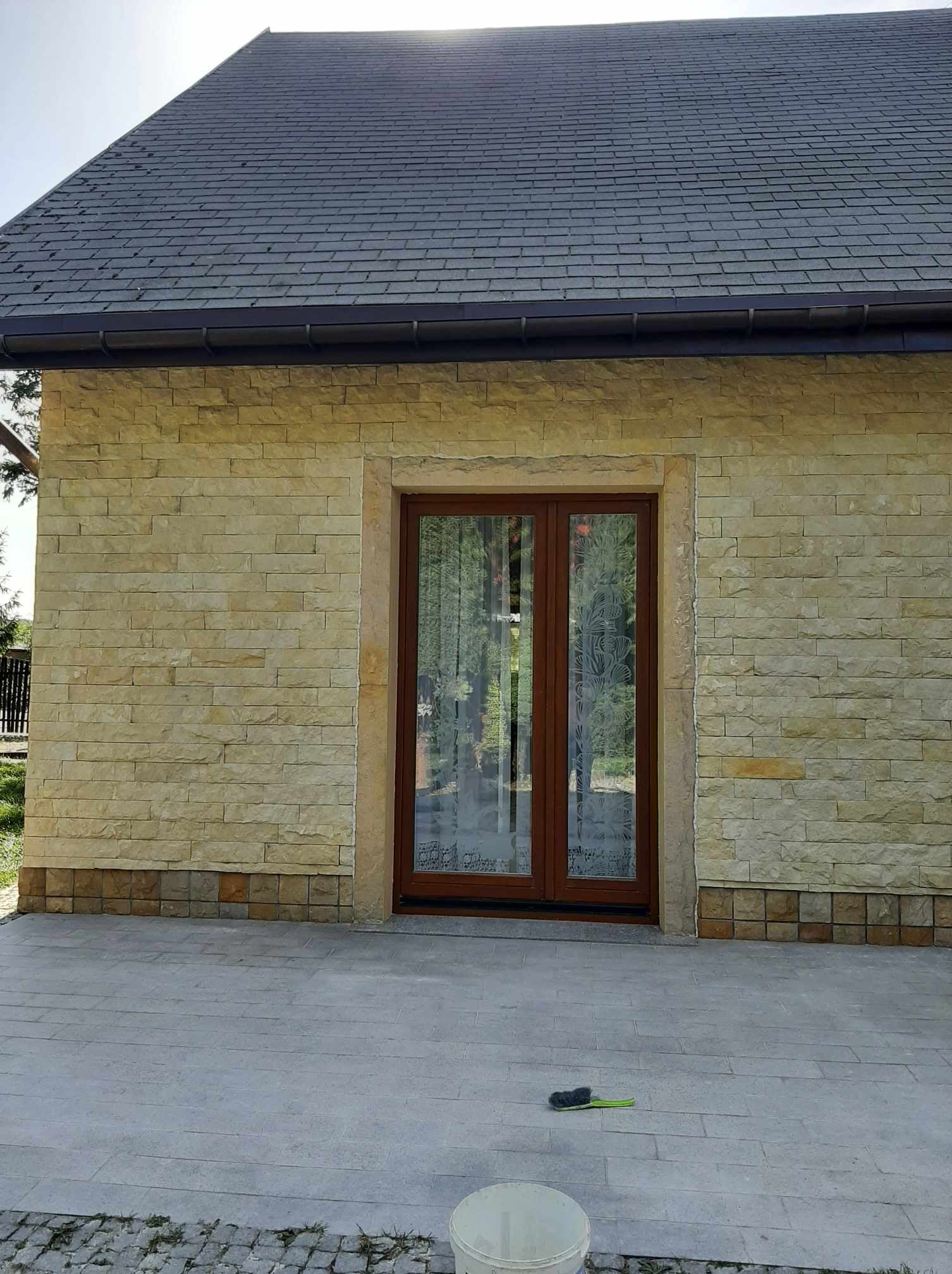 Elewacja budynku z piaskowca z brązowymi drzwiami dwuskrzydłowymi, szary dach z rynną, kostka brukowa na podejściu, widoczny pędzel do fugowania oraz wiadro po farbie.