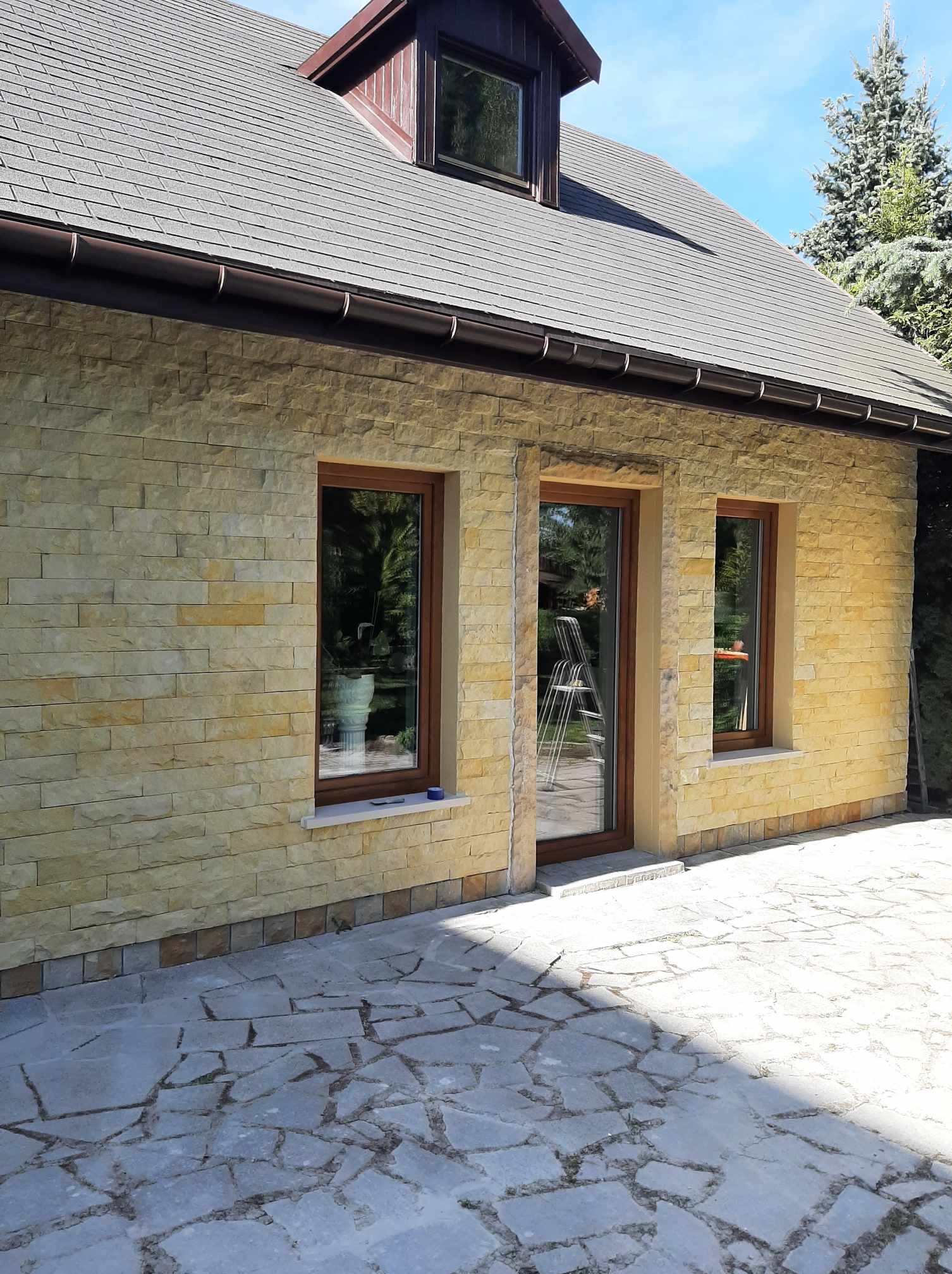 Elewacja domu z piaskowca z trzema oknami w brązowych ramach, widoczny fragment dachu i podwórka z nieregularną kamienną nawierzchnią.