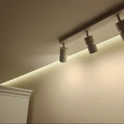 Nowoczesne oświetlenie sufitowe z trzema reflektorami punktowymi i ukrytym paskiem LED, oświetlające gładką, beżową ścianę i fragment białego mebla z dekoracyjnym wykończeniem.