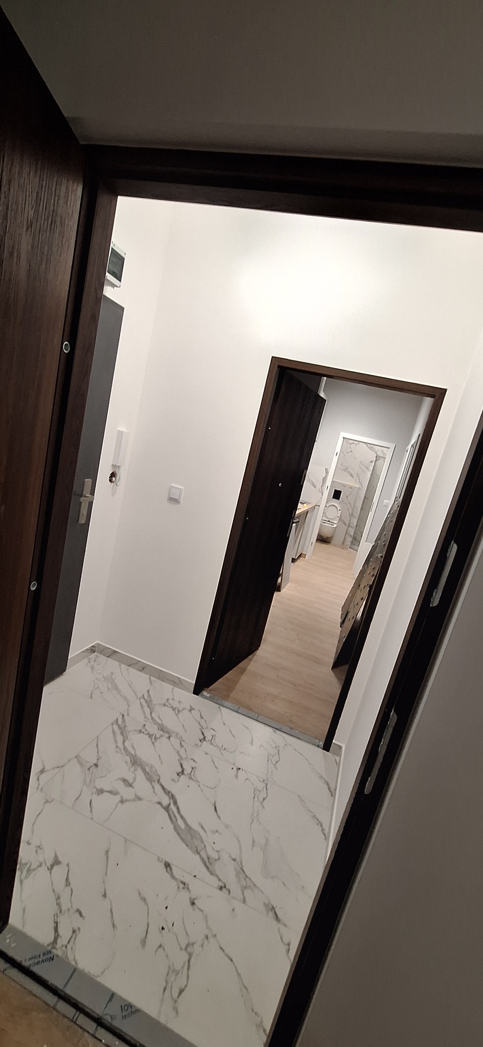 Widok z korytarza na wykończone wnętrze mieszkania z białymi ścianami, marmurową podłogą i drewnianymi drzwiami, w tle widoczna łazienka z białymi płytkami.