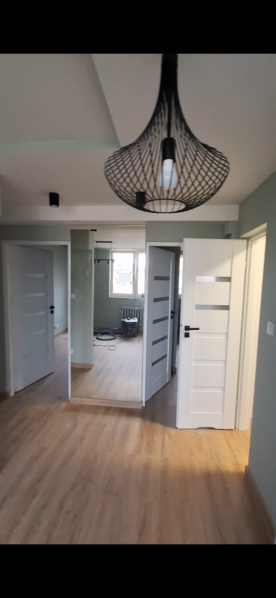 Wnętrze mieszkania w trakcie prac wykończeniowych: widoczne nowe drzwi wewnętrzne w kolorze białym z czarnymi klamkami, podłoga z paneli, surowe ściany w odcieniach szarości i zieleni, oraz ozdobna...