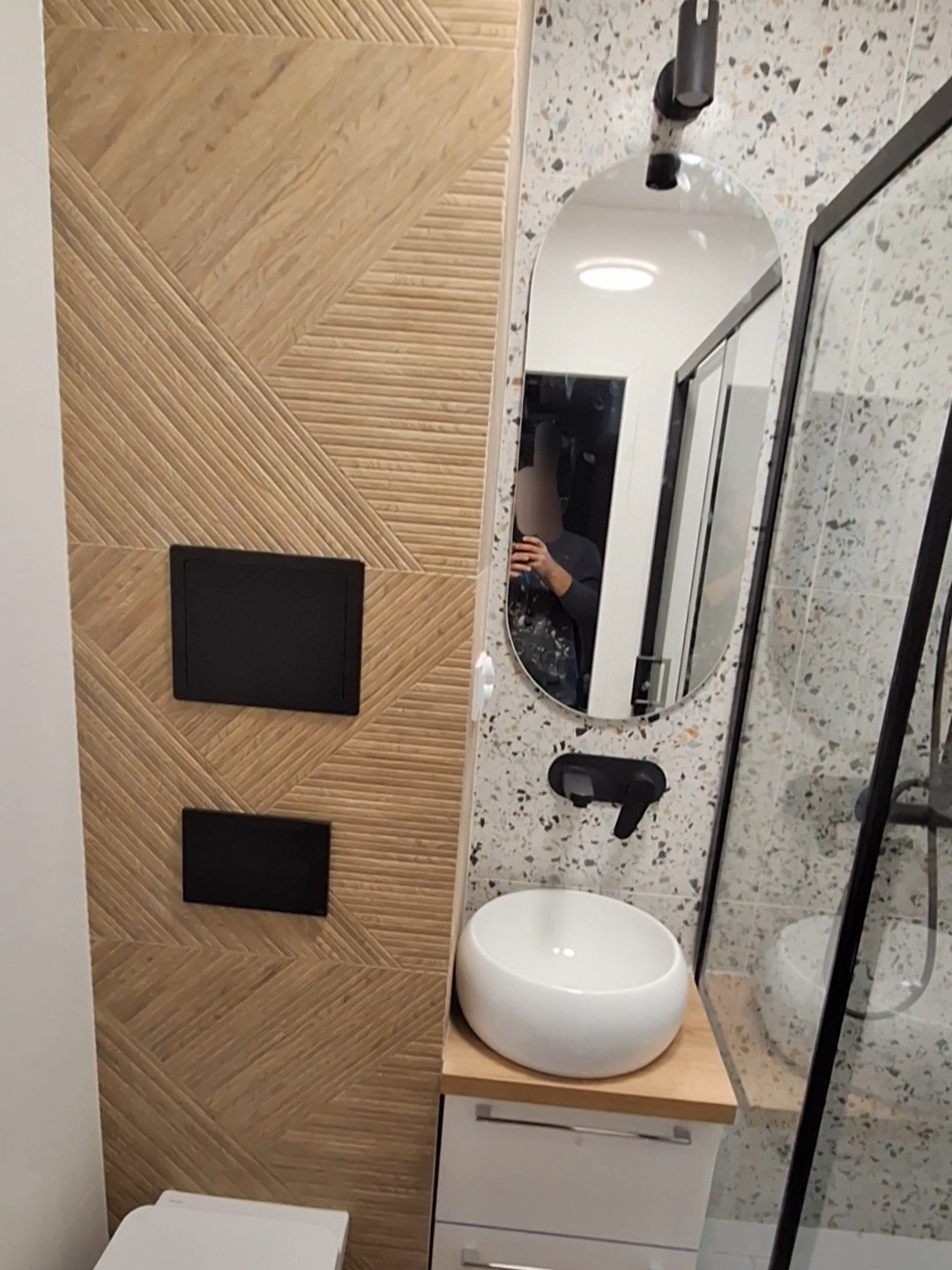 Nowoczesna łazienka z umywalką nablatową, owalnym lustrem z czarną armaturą, ścianą wykończoną płytkami imitującymi drewno oraz kabiną prysznicową ze szklaną ścianką i płytkami lastryko.