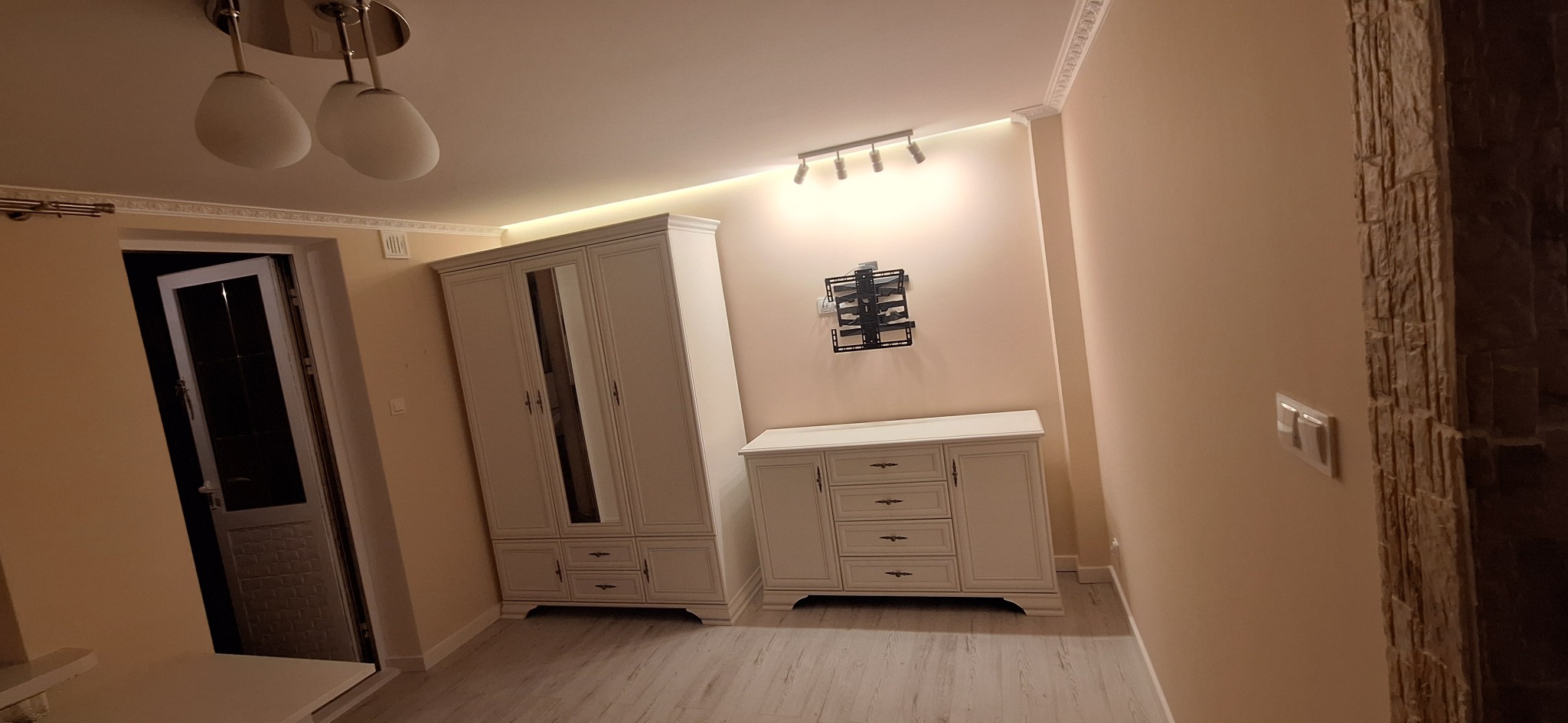 Wykończone wnętrze pokoju z białymi meblami w stylu klasycznym: szafa trzydrzwiowa z lustrem, komoda, oświetlenie punktowe na suficie, uchwyt na telewizor na ścianie i dekoracyjna okładzina...