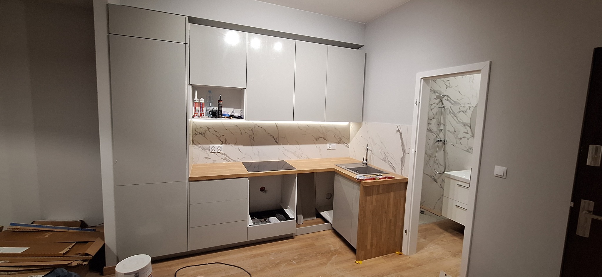 Częściowo ukończona kuchnia z szarymi szafkami, drewnianym blatem i marmurowym backsplashiem, widok na łazienkę z marmurowymi płytkami w tle, na podłodze widoczne materiały budowlane.