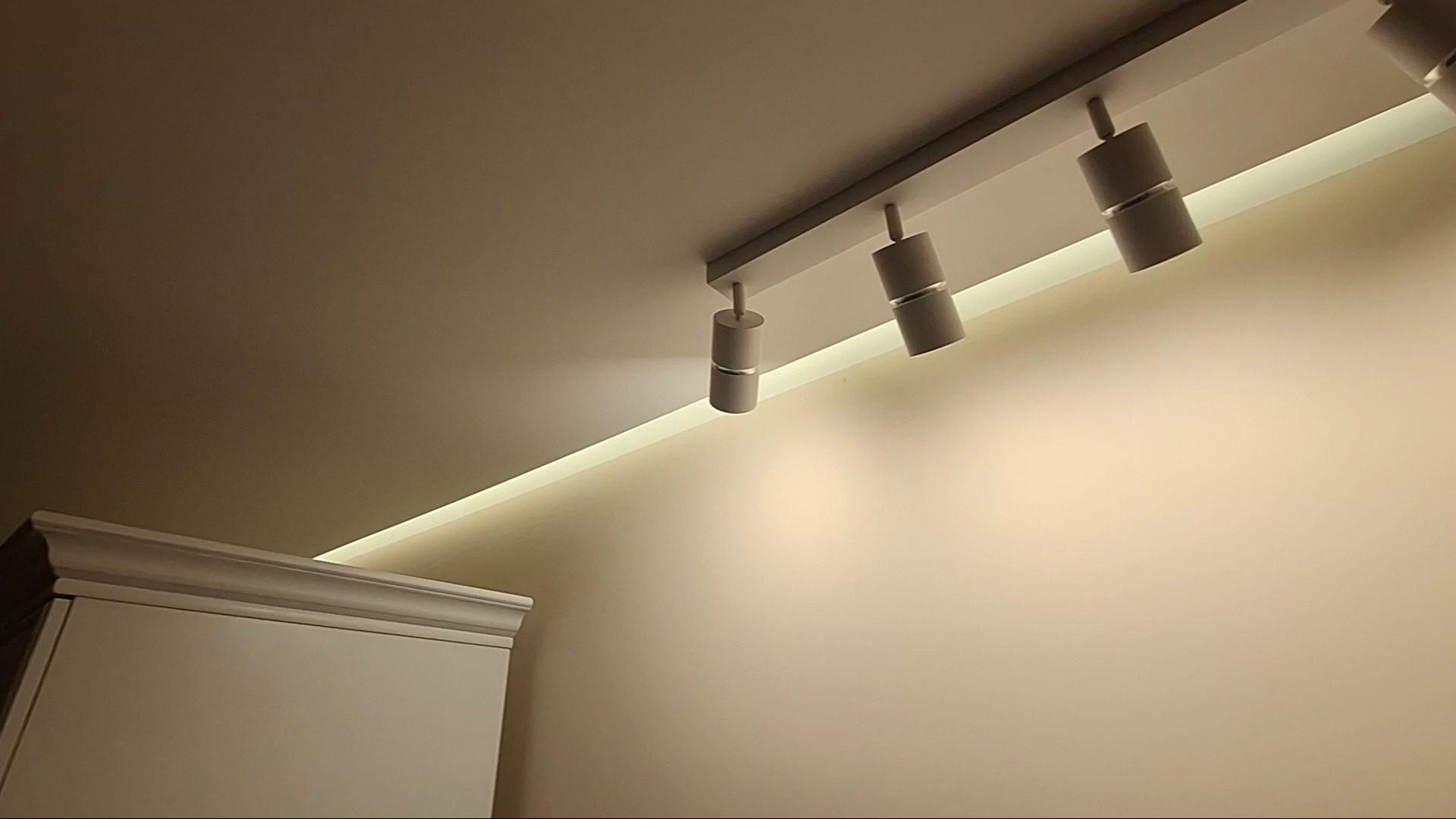 Nowoczesne oświetlenie sufitowe z trzema reflektorami punktowymi i ukrytym paskiem LED, oświetlające gładką, beżową ścianę i fragment białego mebla z dekoracyjnym wykończeniem.