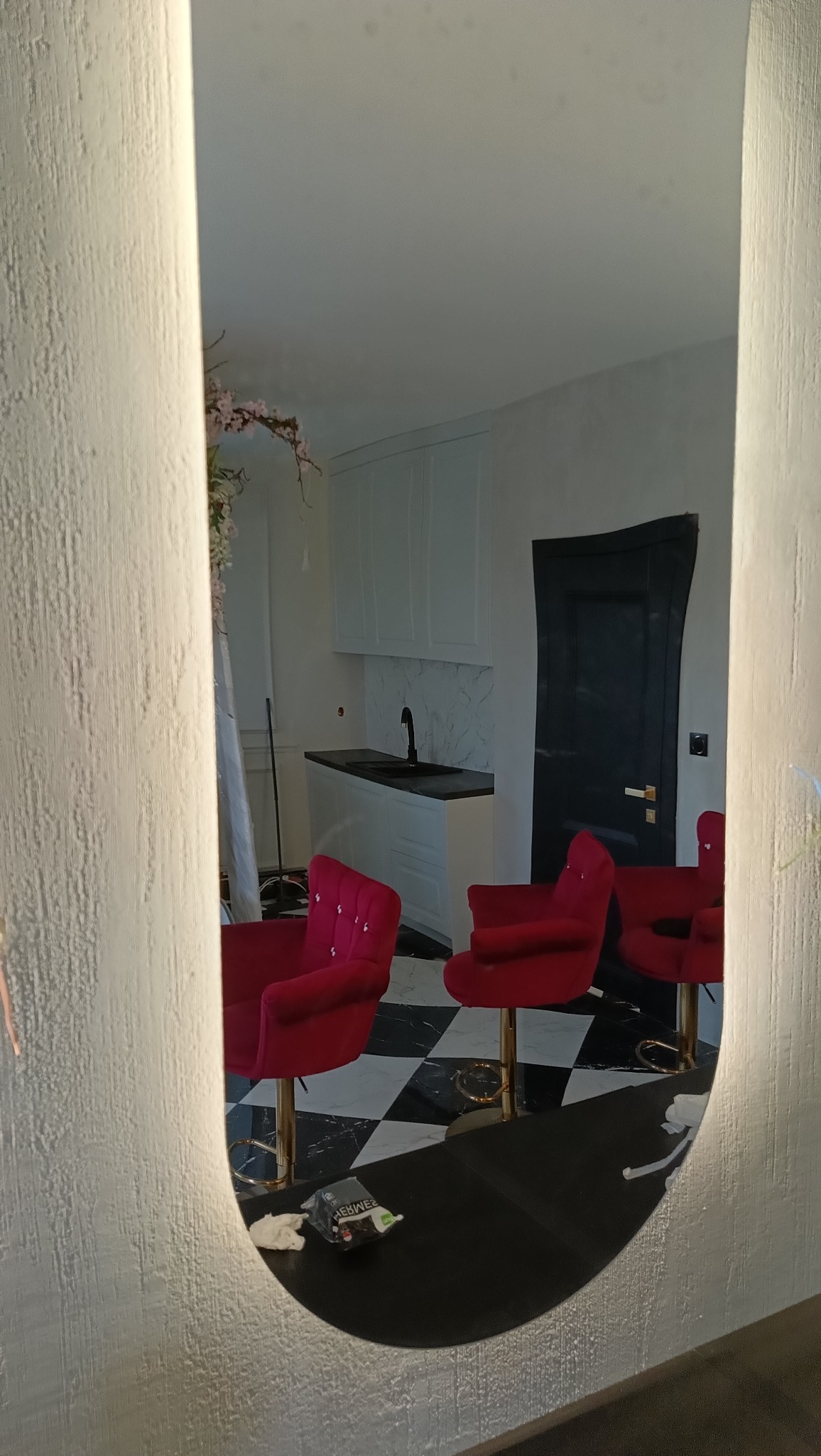 Odbicie w podświetlanym lustrze: wnętrze z białymi szafkami kuchennymi, czarnym blatem, marmurowym backsplash, czarnymi drzwiami i trzema czerwonymi krzesłami barowymi na czarno-białej podłodze.