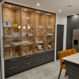 Wysoka przeszklona szafa w zabudowie z szufladami w dolnej części, podświetlona, wypełniona szkłem, porcelaną i dekoracjami, w nowoczesnej kuchni z szarymi frontami i wyspą.