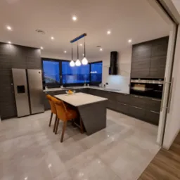 Nowoczesna kuchnia z ciemnymi, drewnopodobnymi szafkami, wyspą z marmurowym blatem i pomarańczowymi krzesłami, oświetlona wiszącymi lampami i punktowymi światłami.