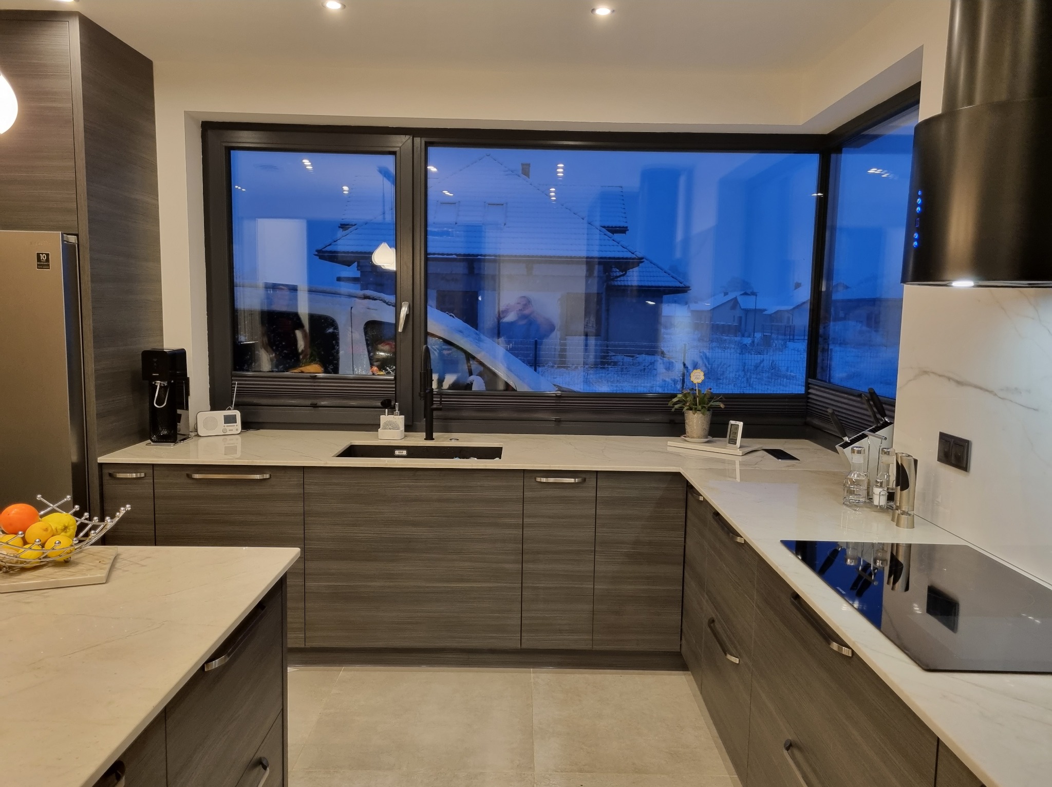 Nowoczesna kuchnia z ciemnymi drewnianymi szafkami, jasnym blatem i dużym oknem z widokiem na zasypany śniegiem dom.