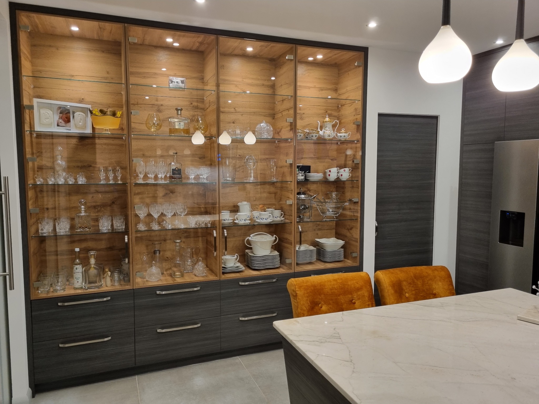 Duża, przeszklona szafa z drewnianym wnętrzem i szarymi szufladami w nowoczesnej kuchni, oświetlona punktowymi światłami, eksponująca kolekcję szkła, porcelany i pamiątek.